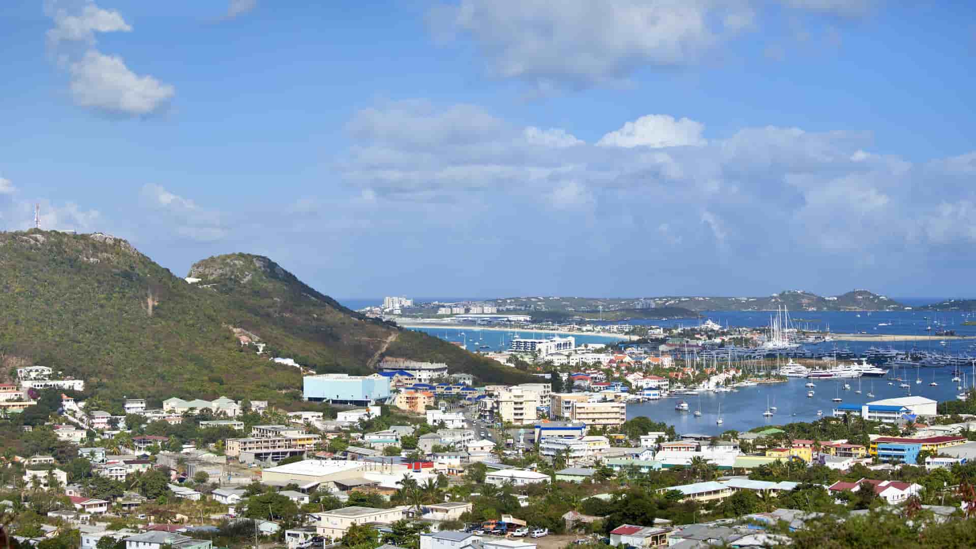 A high-angle view of the bustling harbor of Marigot, St. Martin, with boats and yachts docked along the coast and colorful buildings nestled against the green hills.