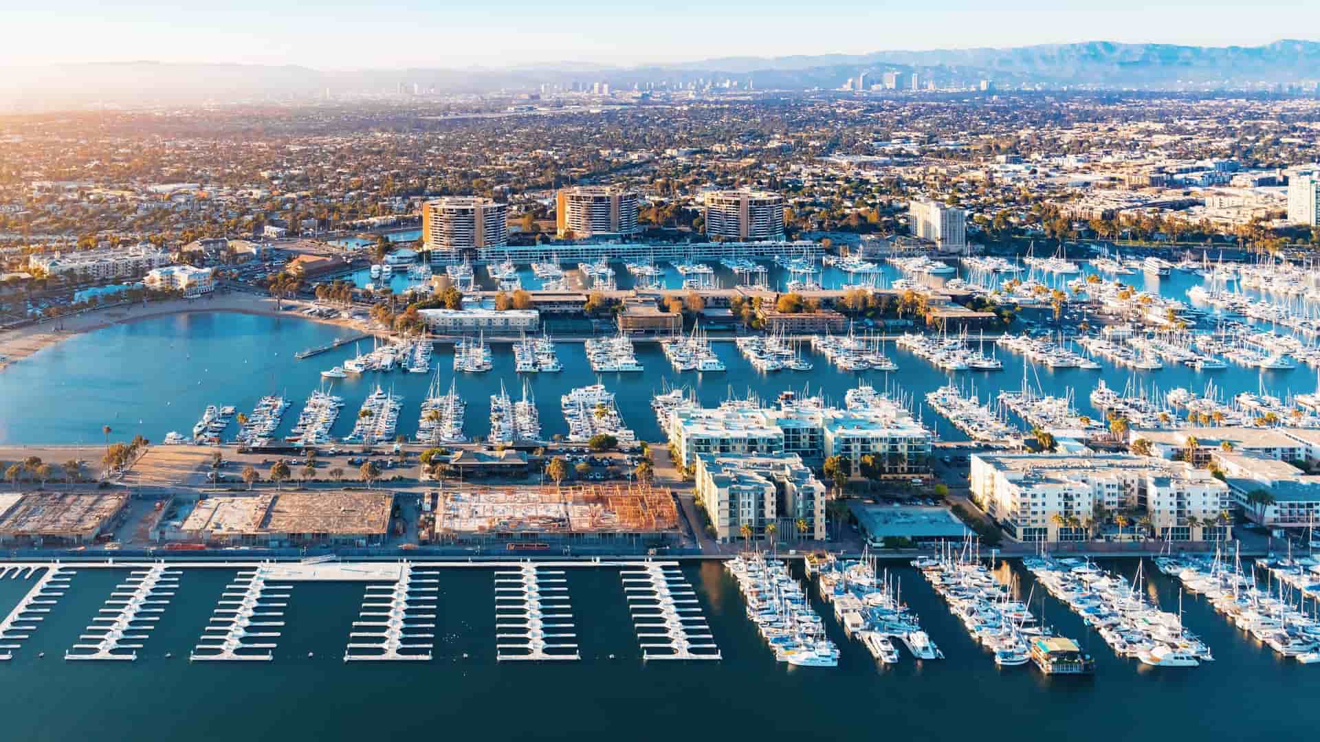 An aerial view of the Marina del Rey seaside community in Los Angeles, California. The image captures the man-made harbor filled with boats and yachts, surrounded by high-rise apartment buildings, commercial developments, and recreational areas, with the Pacific Ocean visible to the left.