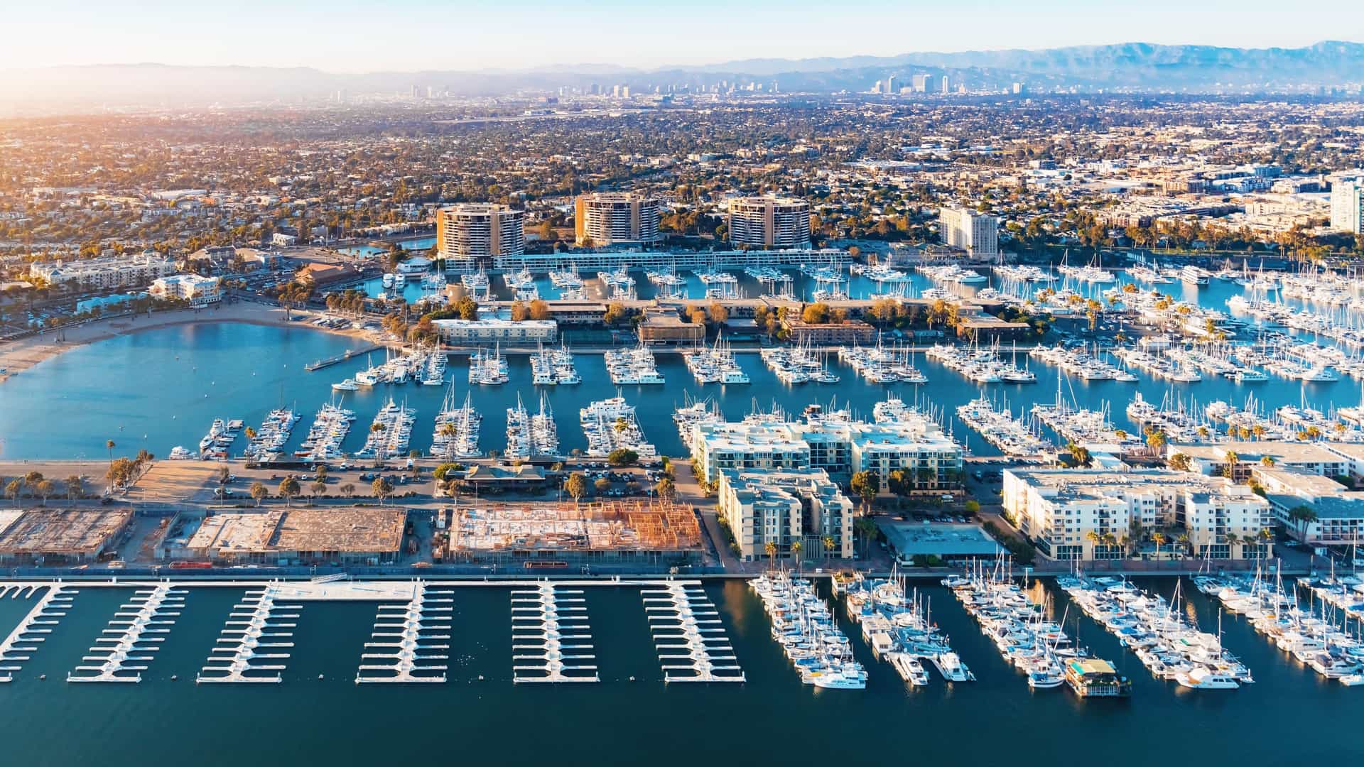 An aerial view of the Marina del Rey seaside community in Los Angeles, California. The image captures the man-made harbor filled with boats and yachts, surrounded by high-rise apartment buildings, commercial developments, and recreational areas, with the Pacific Ocean visible to the left.