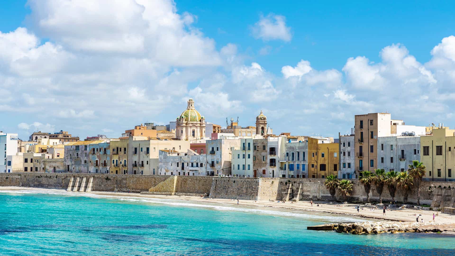 "A coastal view of the city of Marsala, Italy, with historic buildings and a prominent cathedral dome overlooking the turquoise Mediterranean Sea on a sunny day. "