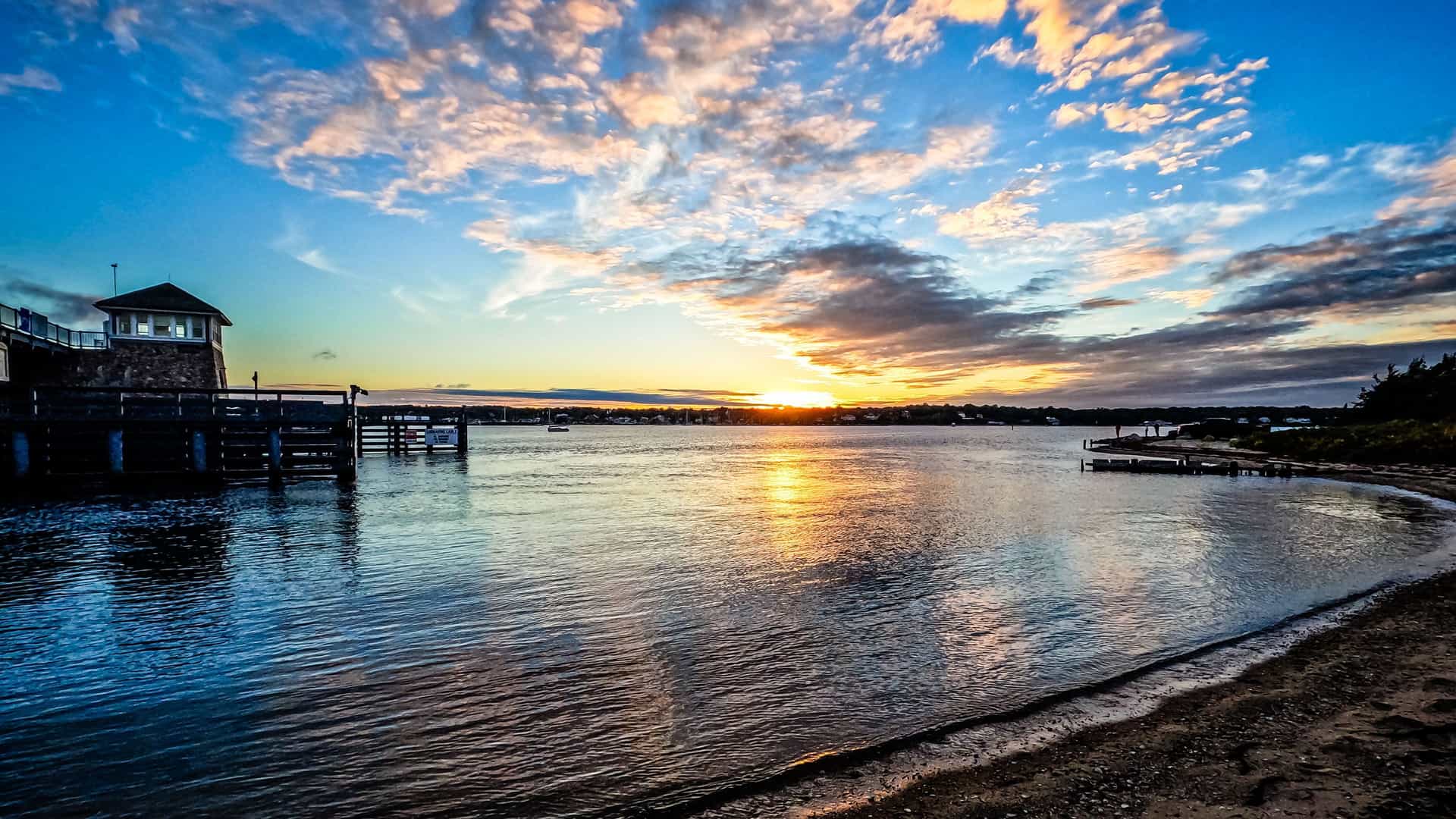 A scenic sunset over the calm waters of Martha's Vineyard, with the sun setting on the horizon and reflecting on the water near a small dock and sandy beach.