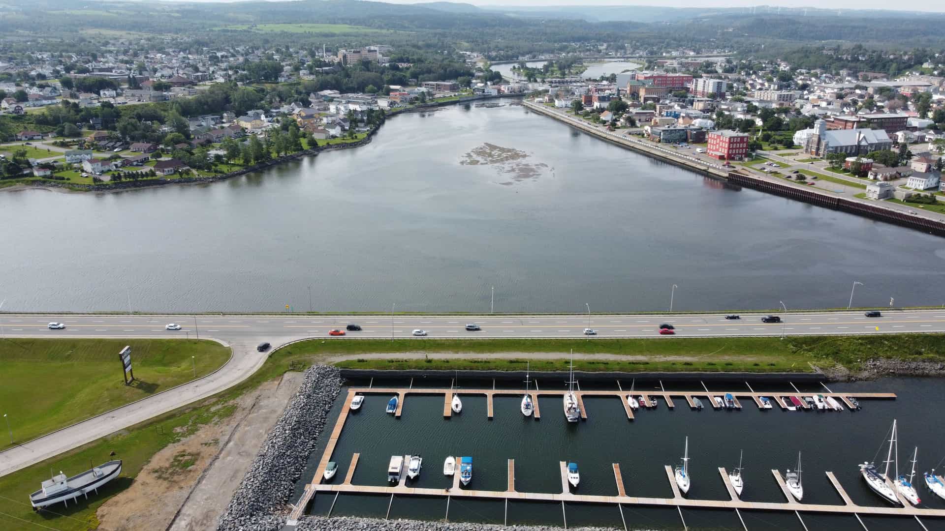 "An aerial view of the city of Matane, Canada, showing the river and marina with boats, a highway, and the town's buildings and rolling hills in the background. "