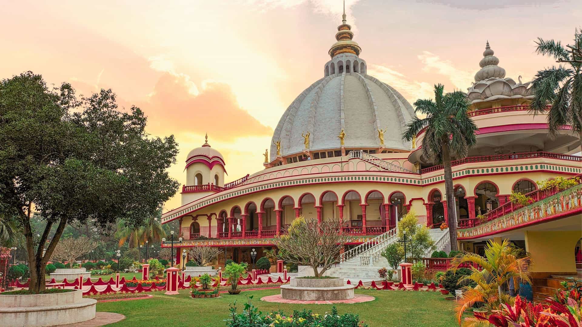A grand view of the ISKCON temple in Mayapur, India, a significant pilgrimage site with a large, white dome and ornate architecture surrounded by well-manicured gardens.