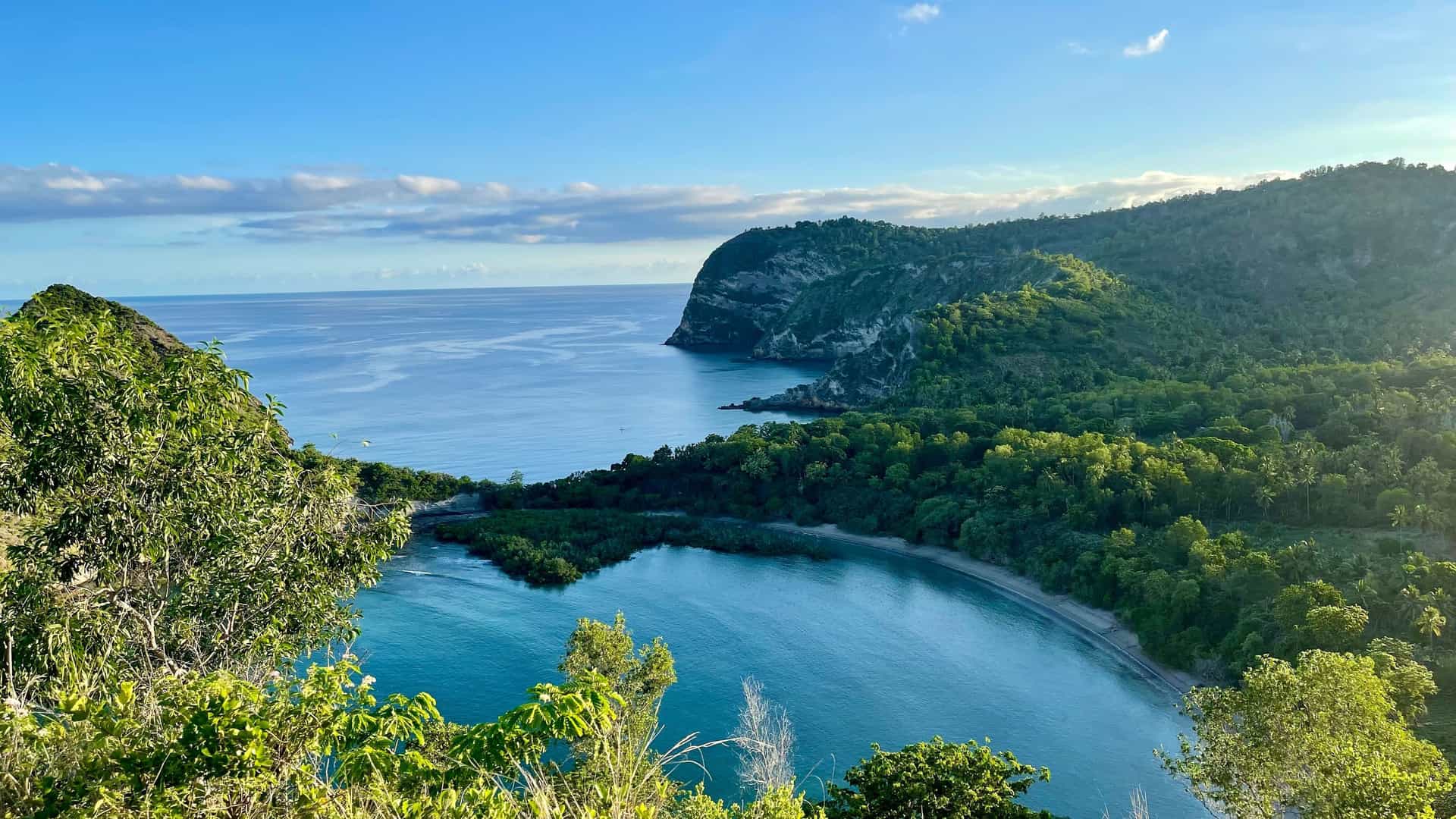 A breathtaking high-angle view of a serene bay in Mayotte, with lush green hills surrounding the turquoise water and a cliff face meeting the open ocean.