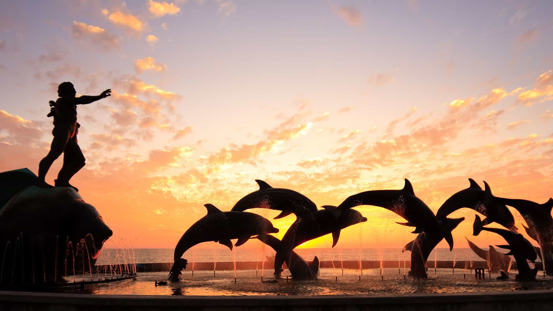 A breathtaking sunset view of the "Monumento al Pescador" fountain in Mazatlán, Mexico, with a silhouette of a man and dolphins against an orange and pink sky.
