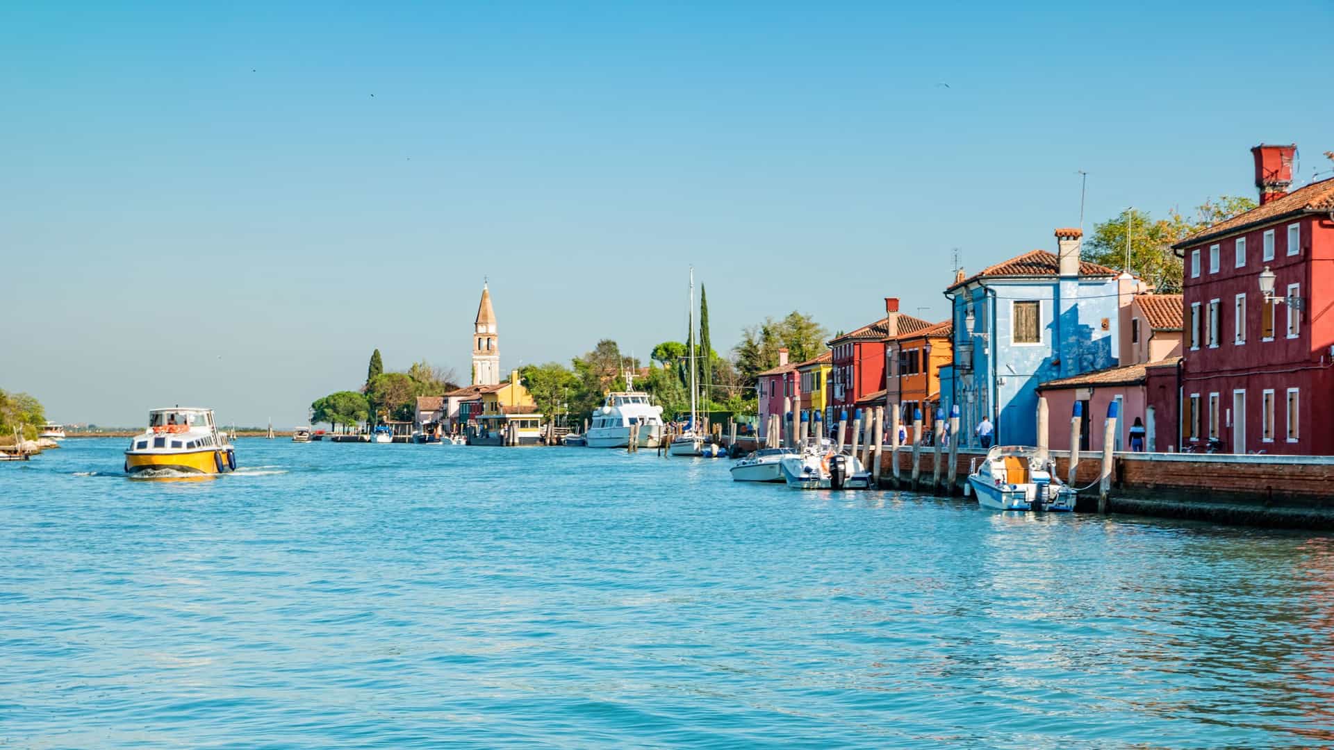 A beautiful canal view on Mazzorbo Island, with colorful buildings lining the water, docked boats, and a church tower visible in the distance.