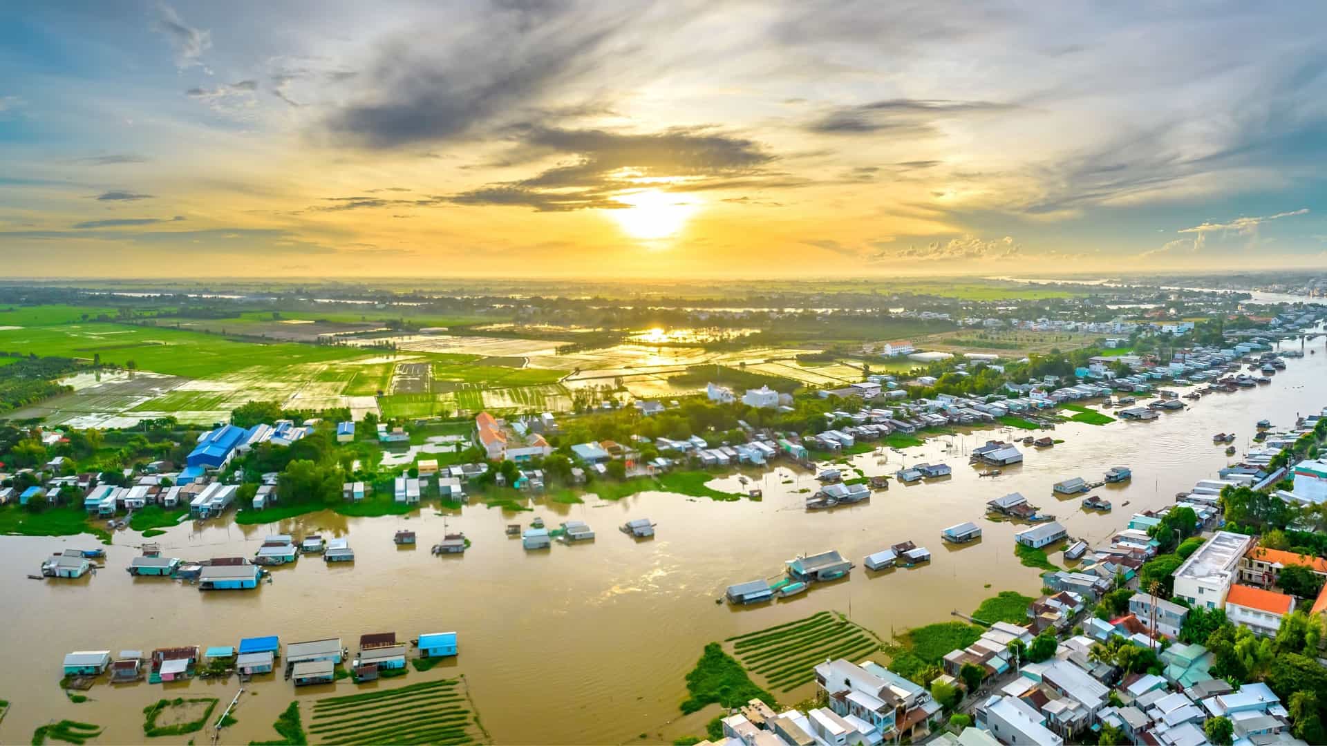 An aerial sunset view of the floating houses on the Mekong River in Vietnam, with vast green rice fields and a vibrant sky in the background.