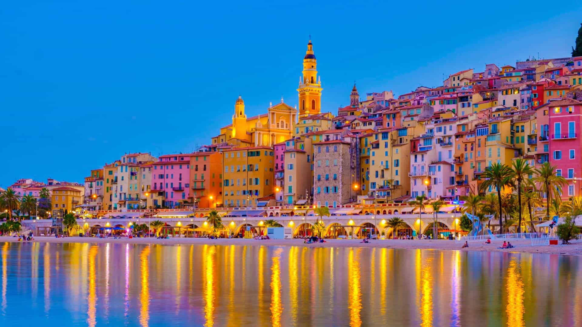 A breathtaking dusk shot of the colorful coastal town of Menton, France, with its vibrant buildings and a prominent church tower reflecting on the still water of the Mediterranean Sea.
