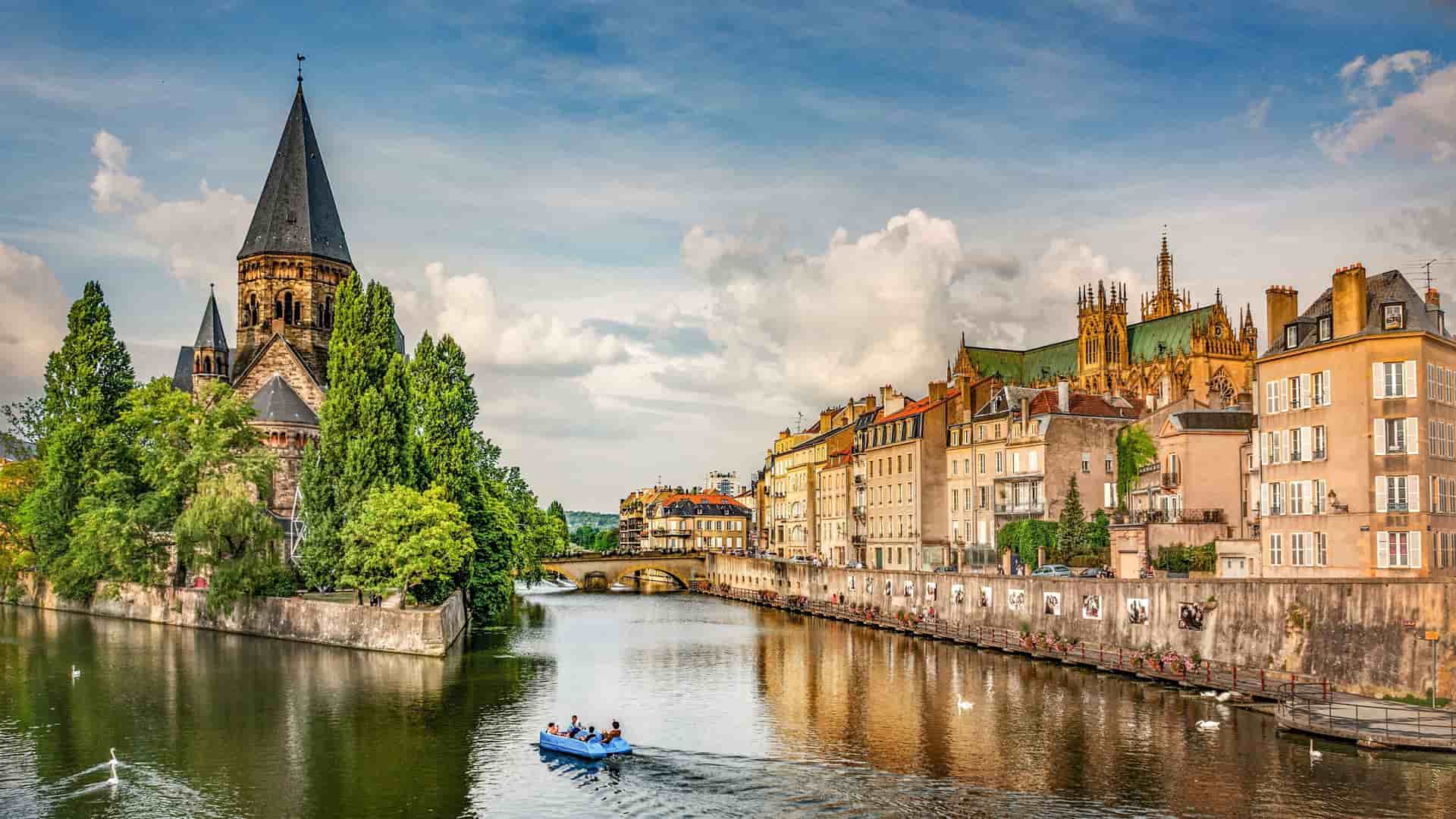 A picturesque view of the city of Metz, France, with a person paddling a small boat on the Moselle River. The scene features the Temple Neuf and colorful buildings along the waterfront.