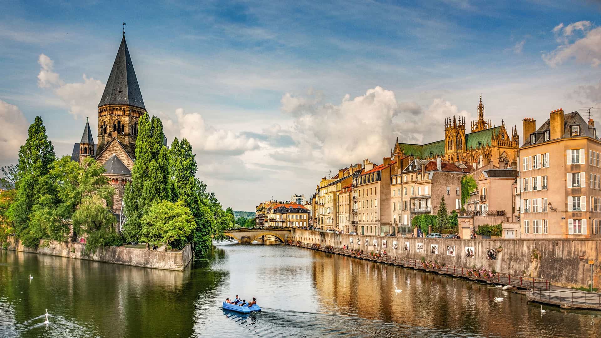 A picturesque view of the city of Metz, France, with a person paddling a small boat on the Moselle River. The scene features the Temple Neuf and colorful buildings along the waterfront.