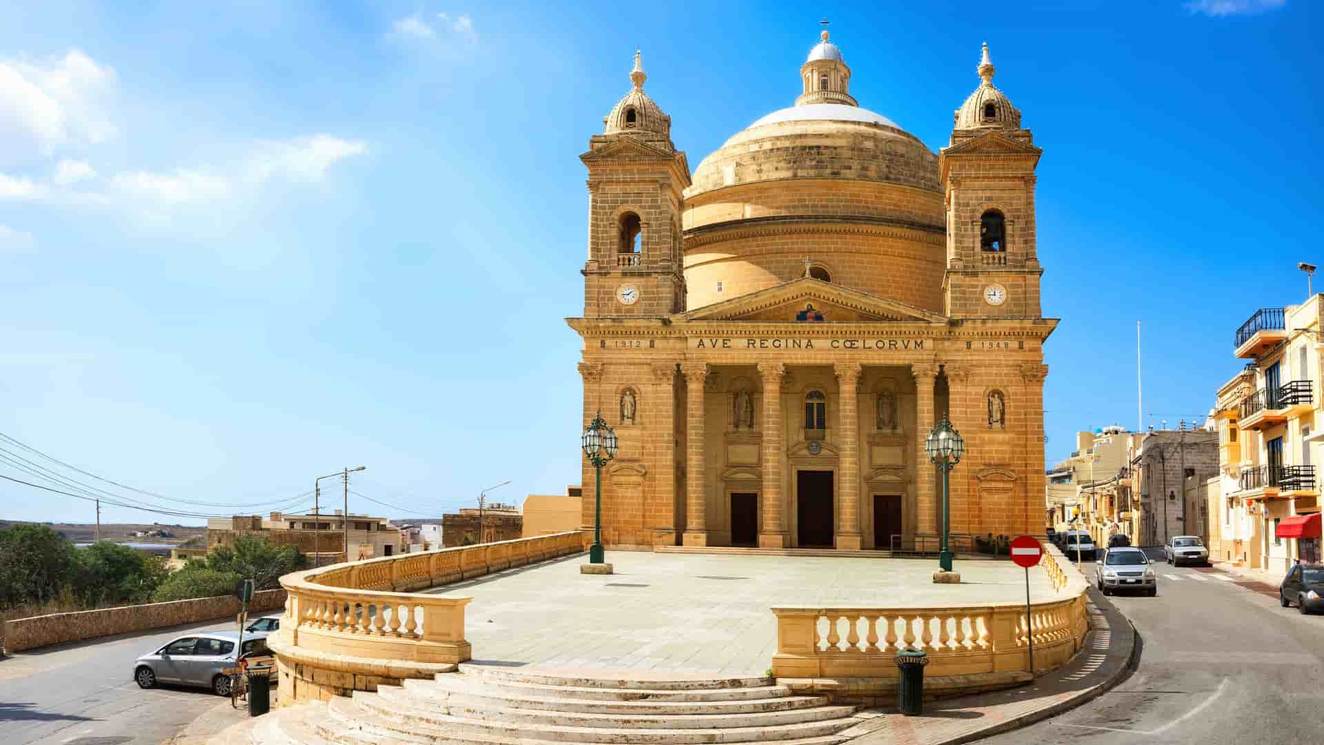 A stunning low-angle shot of the Parish Church of Saint Mary of Mġarr, a monumental stone temple with a large dome and two bell towers, located on the island of Gozo in Malta.