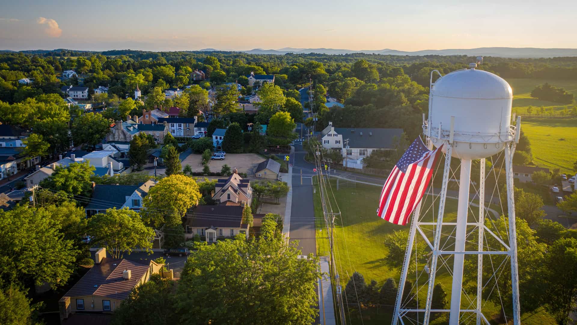 A warm sunset aerial view in Middleburg featuring a tall white water tower draped with an American flag, surrounded by tree‑lined streets, small homes, and rolling green hills glowing in golden light.