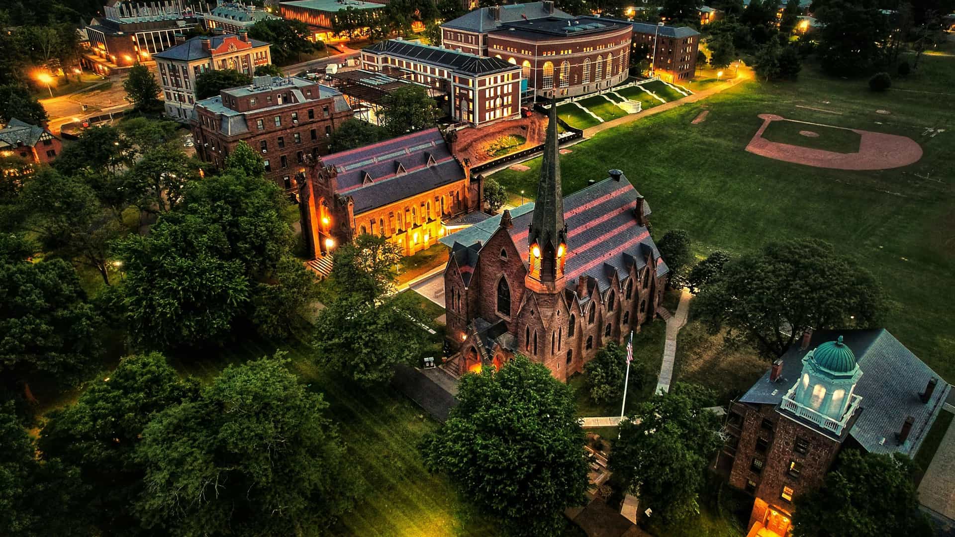 "An aerial dusk view of the Wesleyan University campus in Middletown, Connecticut, with the historic Memorial Chapel and surrounding buildings lit up at night. "