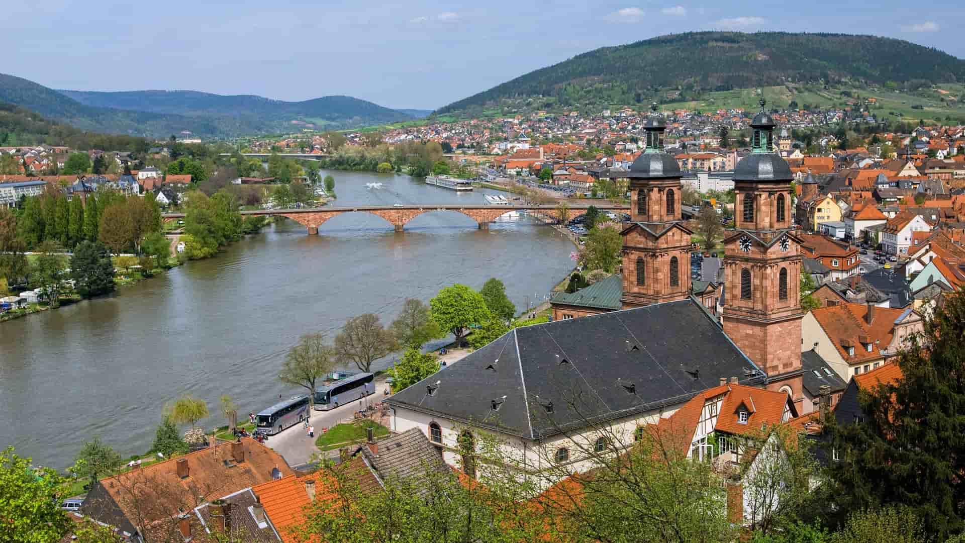 A scenic view of Miltenberg, Germany, with the Main River winding through the town, a prominent church with twin spires in the foreground, and lush green hills in the distance.