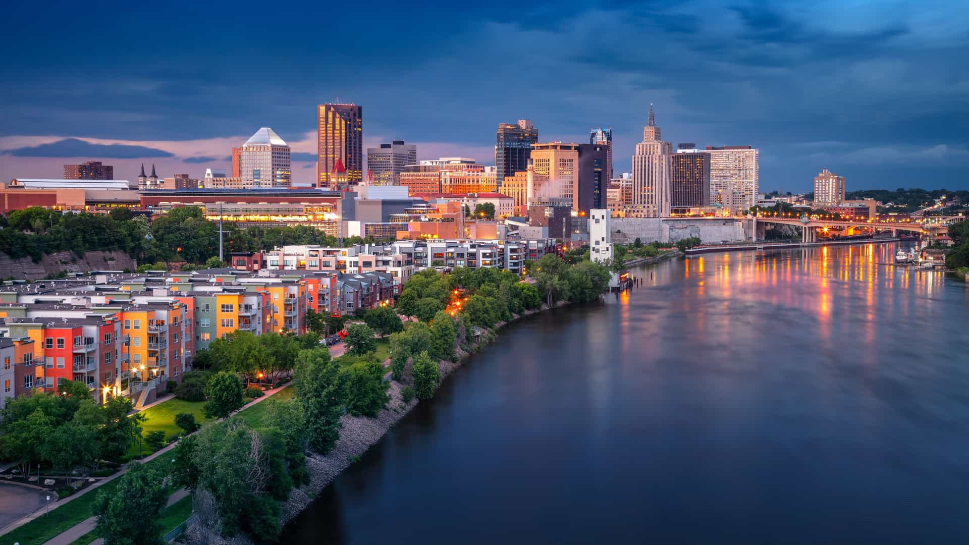 "An aerial night shot of the Minneapolis, Minnesota, skyline, with the Mississippi River reflecting the illuminated downtown buildings, colorful residential apartments, and bridges. "