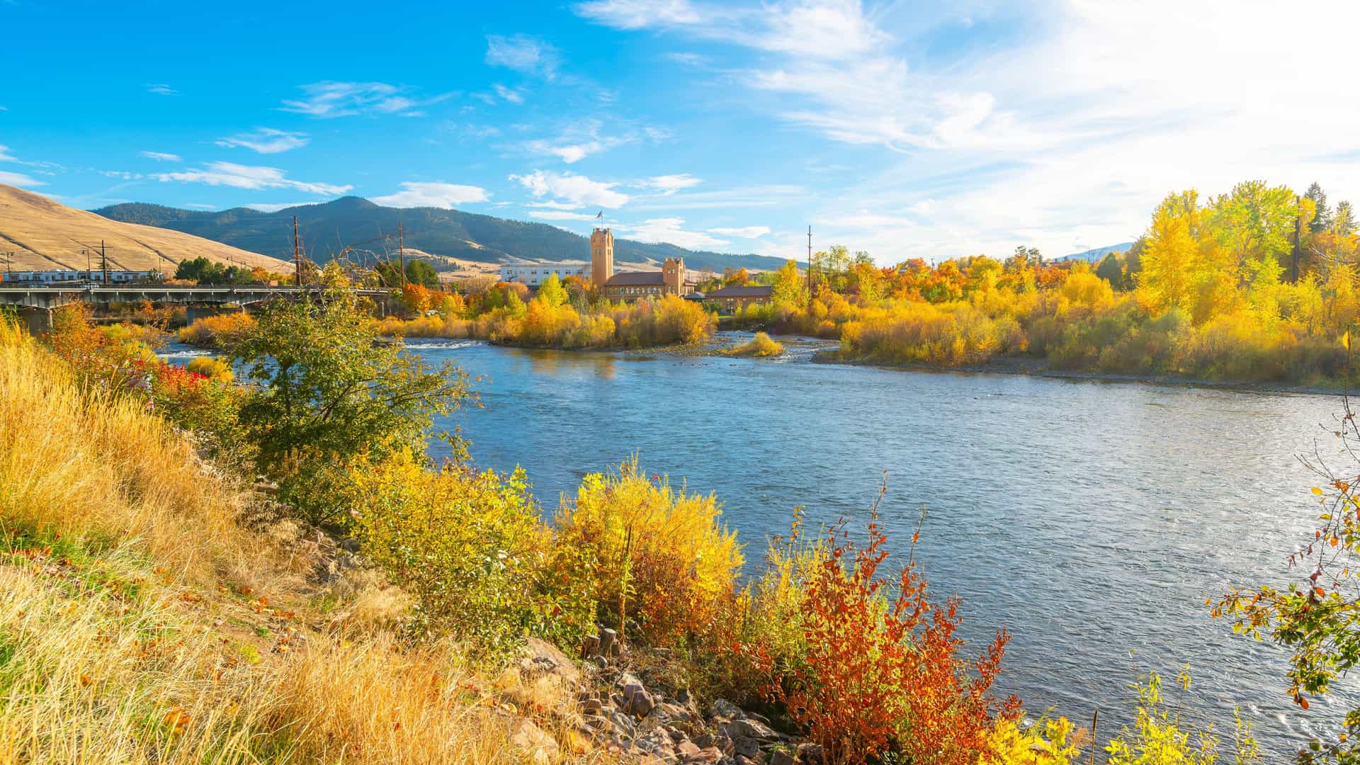 "A stunning autumn view of the Clark Fork River in Missoula, Montana, with the historic Milwaukee Depot in the background and vibrant yellow foliage along the riverbank. "
