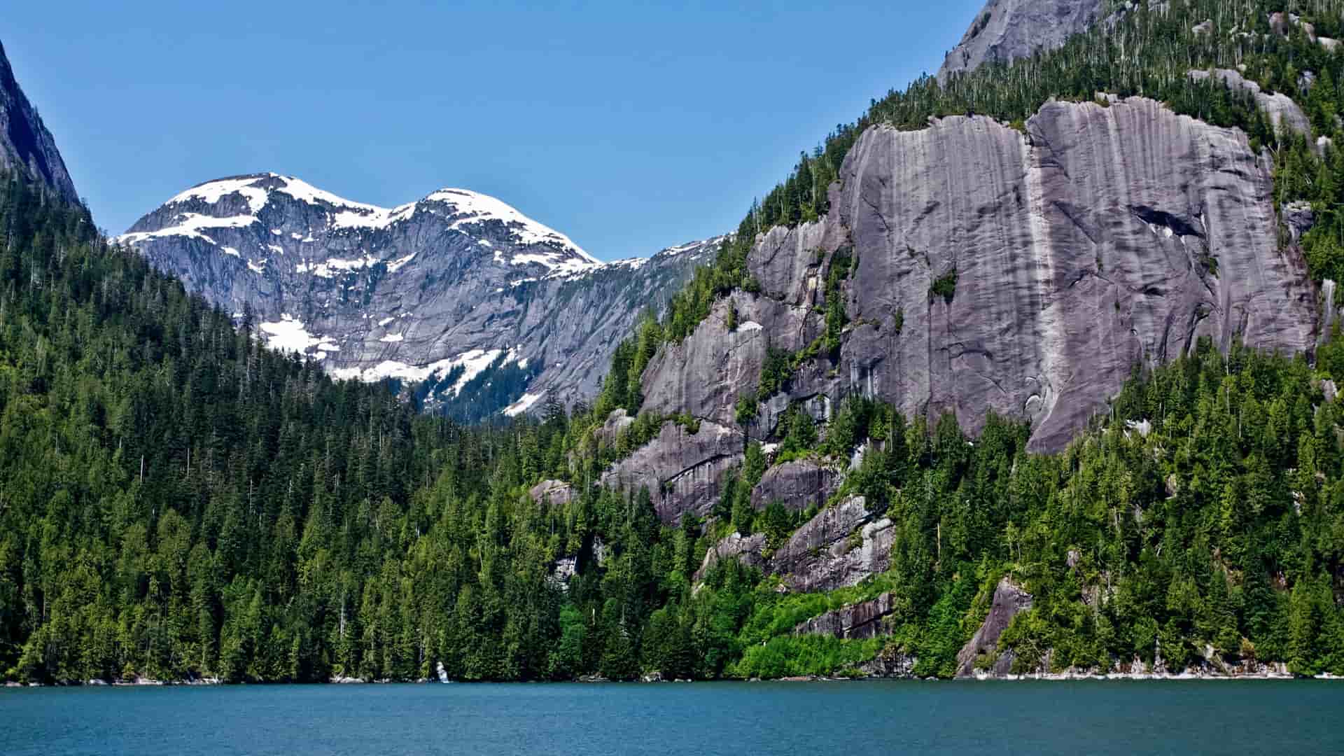 A dramatic view of a pristine fjord in Misty Fjords National Monument, Alaska, with sheer granite cliffs, dense coniferous forests, and snow-capped mountains reflected in the calm water.