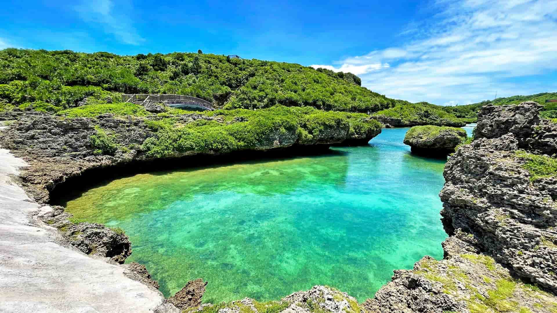 A breathtaking view of an emerald green, natural tidal pool, surrounded by rugged limestone cliffs and lush vegetation, on the island of Miyakojima, Japan.