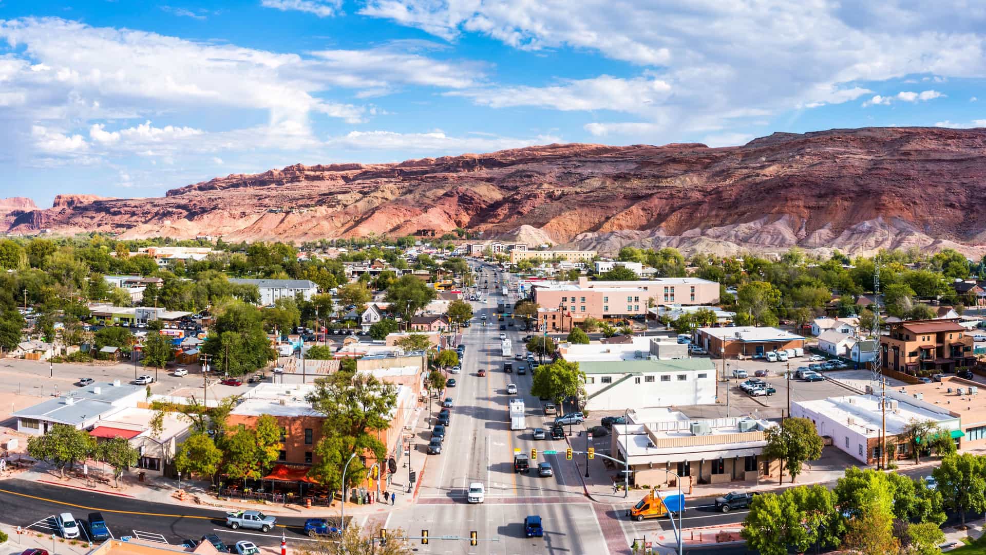 "An aerial panoramic view of downtown Moab, Utah, showing the main street, buildings, and surrounding landscape with the majestic red rock mountains in the background under a blue sky. "