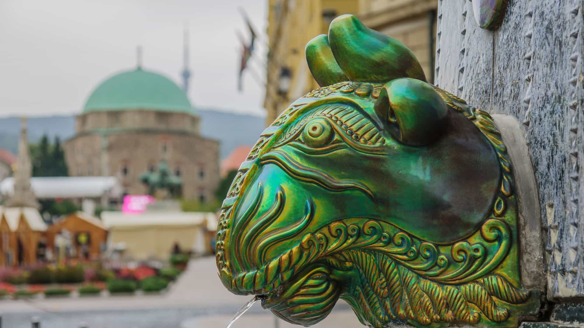 A close-up of a green, ram-like fountain statue in the town of Mohacs, Hungary, with a trickle of water flowing from its mouth and a domed church in the background.