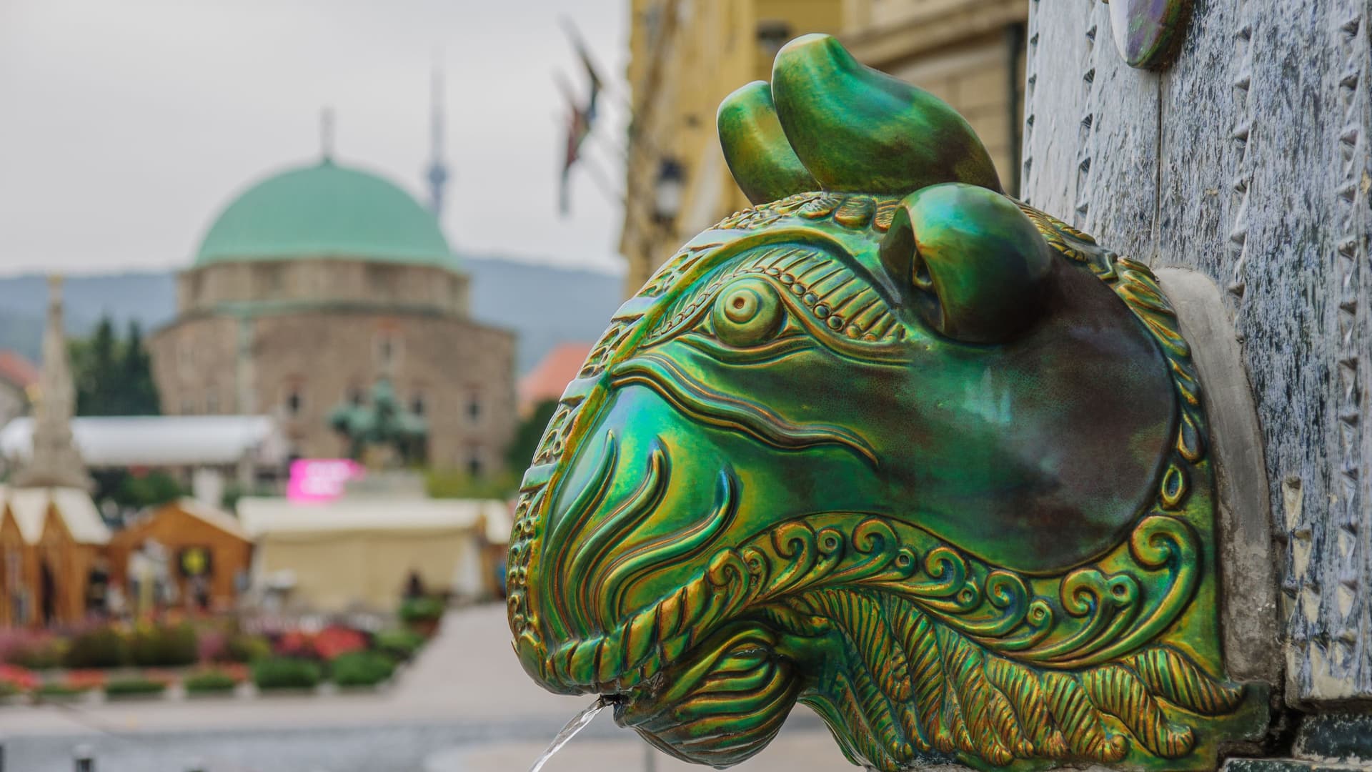 A close-up of a green, ram-like fountain statue in the town of Mohacs, Hungary, with a trickle of water flowing from its mouth and a domed church in the background.