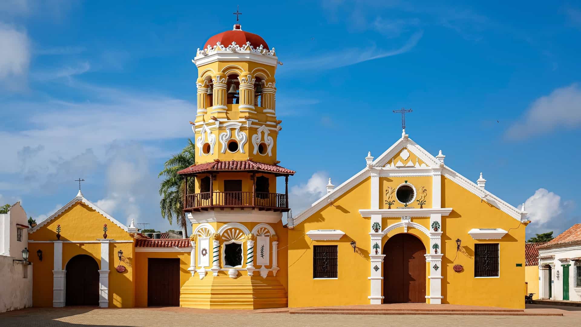 A beautiful colonial-era church in Mompox, Colombia, with its vibrant yellow facade, ornate bell tower, and bright red dome standing out against a clear blue sky.