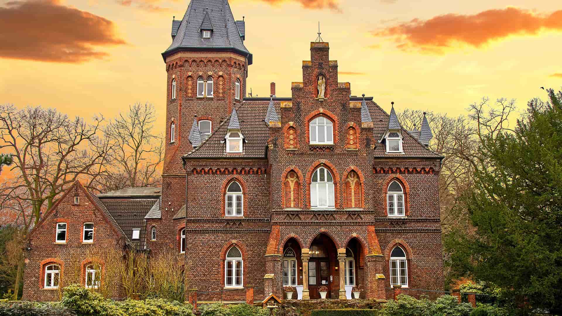 "A front view of the historic Monheim Castle in Monheim am Rhein, Germany, a red brick building with a tower, set against a stunning orange and gold sunset sky. "