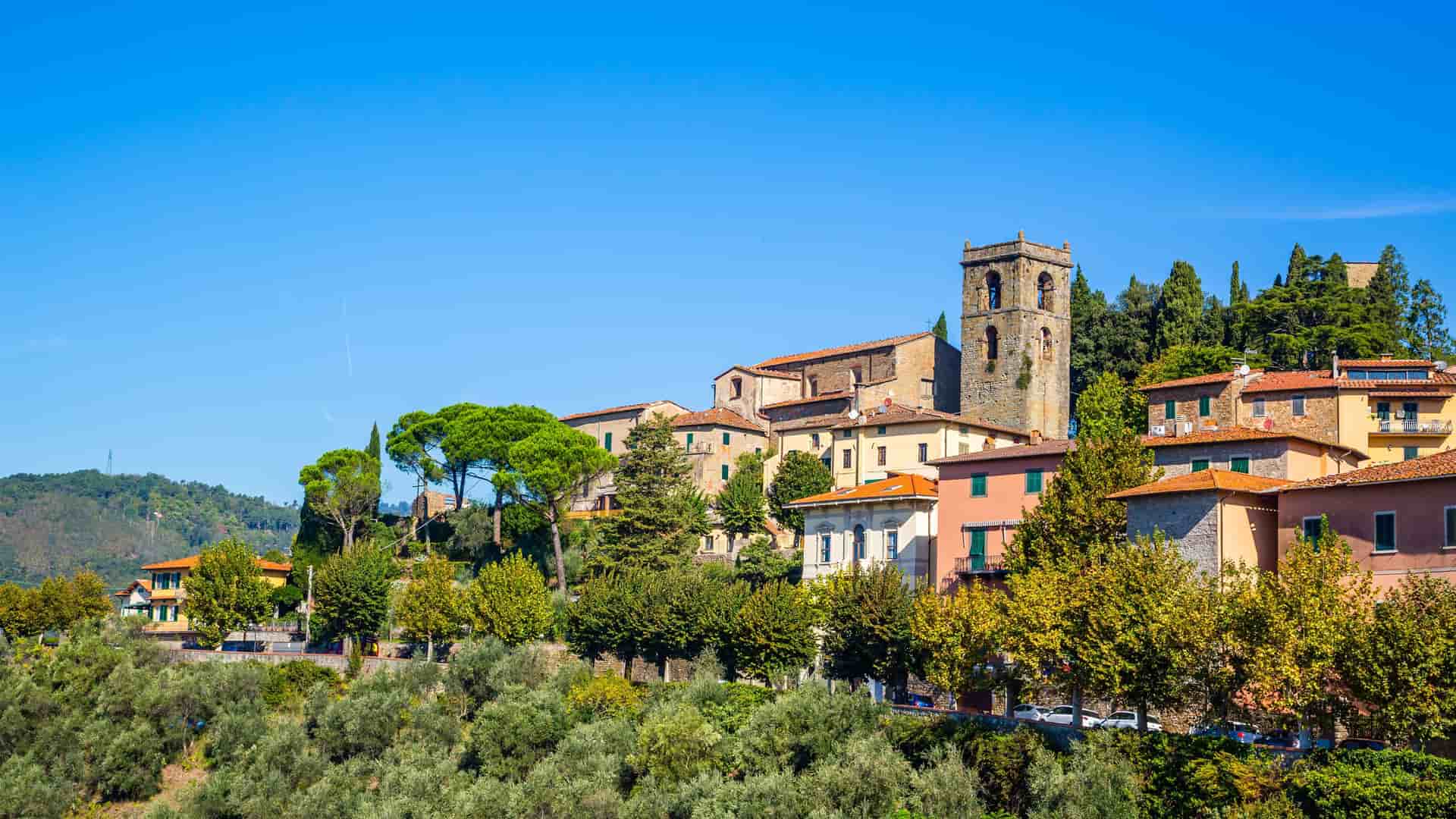 "A beautiful panoramic view of the medieval hilltop village of Montecatini Alto, Italy, with its historic buildings, ancient bell tower, and lush green trees against a clear blue sky. "
