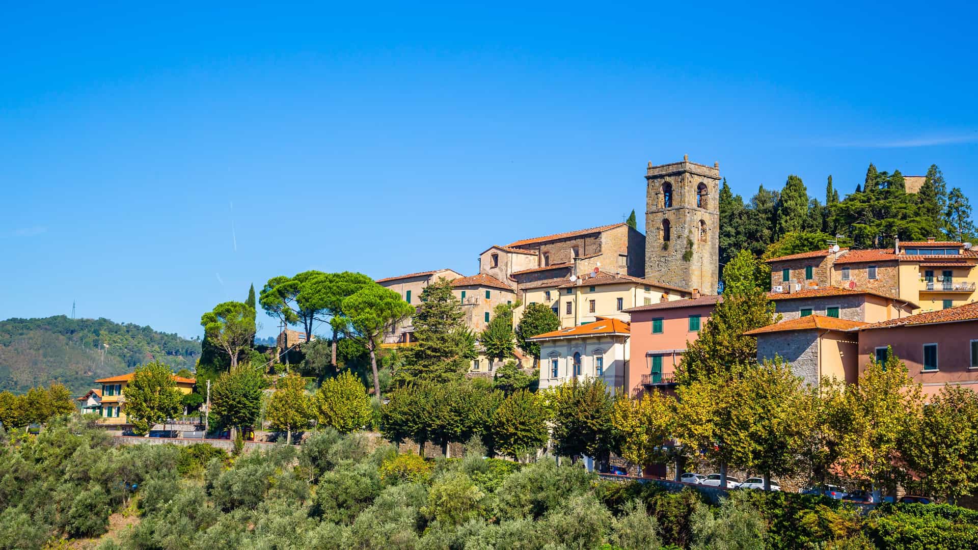 "A beautiful panoramic view of the medieval hilltop village of Montecatini Alto, Italy, with its historic buildings, ancient bell tower, and lush green trees against a clear blue sky. "
