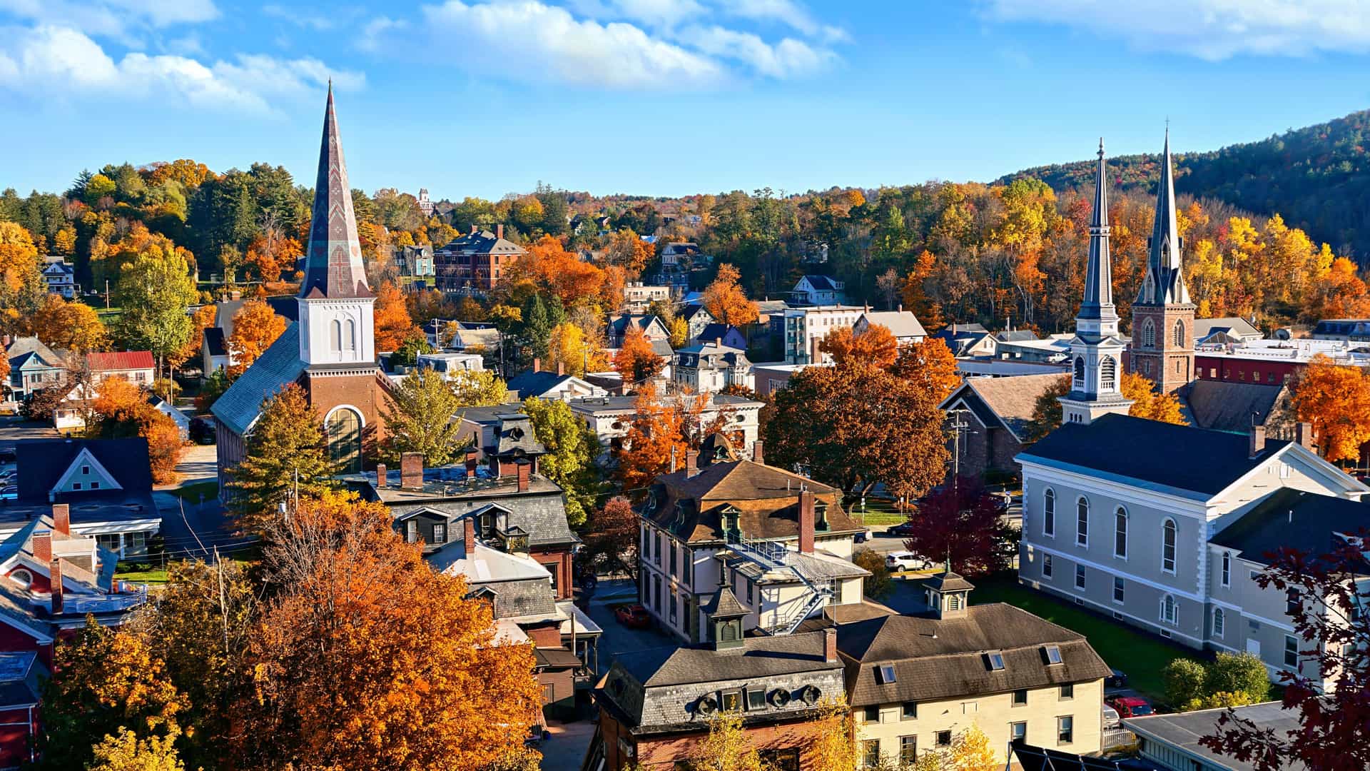 "An aerial autumn view of Montpelier, Vermont, with its historic buildings, two prominent church steeples, and vibrant fall foliage blanketing the surrounding hillsides. "