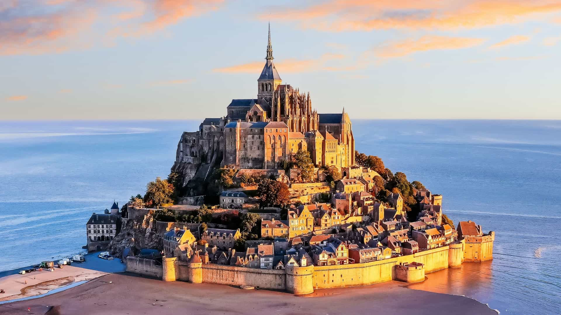 A high-angle view of the iconic Mont Saint-Michel, a tidal island and commune in Normandy, France, with its stunning medieval abbey and village rising from the sea at sunset.