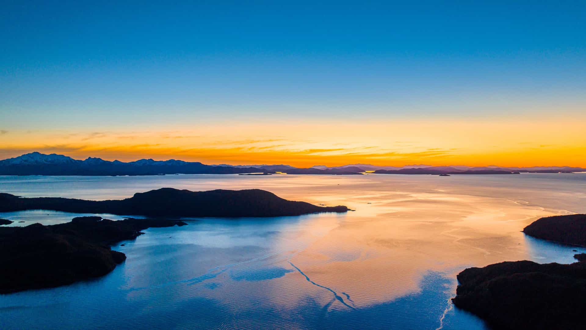 An aerial view of the stunning Moraleda Channel in the Aysén Region of Chile, with dark, rocky islands and tranquil water reflecting the brilliant colors of a sunrise or sunset.