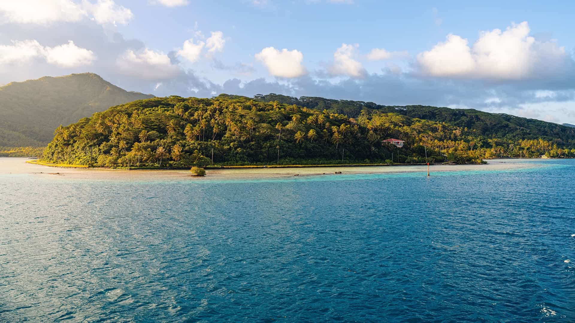A serene view of Motu Mahaea, a small, lush tropical islet in French Polynesia, surrounded by clear turquoise lagoons and the deep blue ocean, with a green mountain in the background.