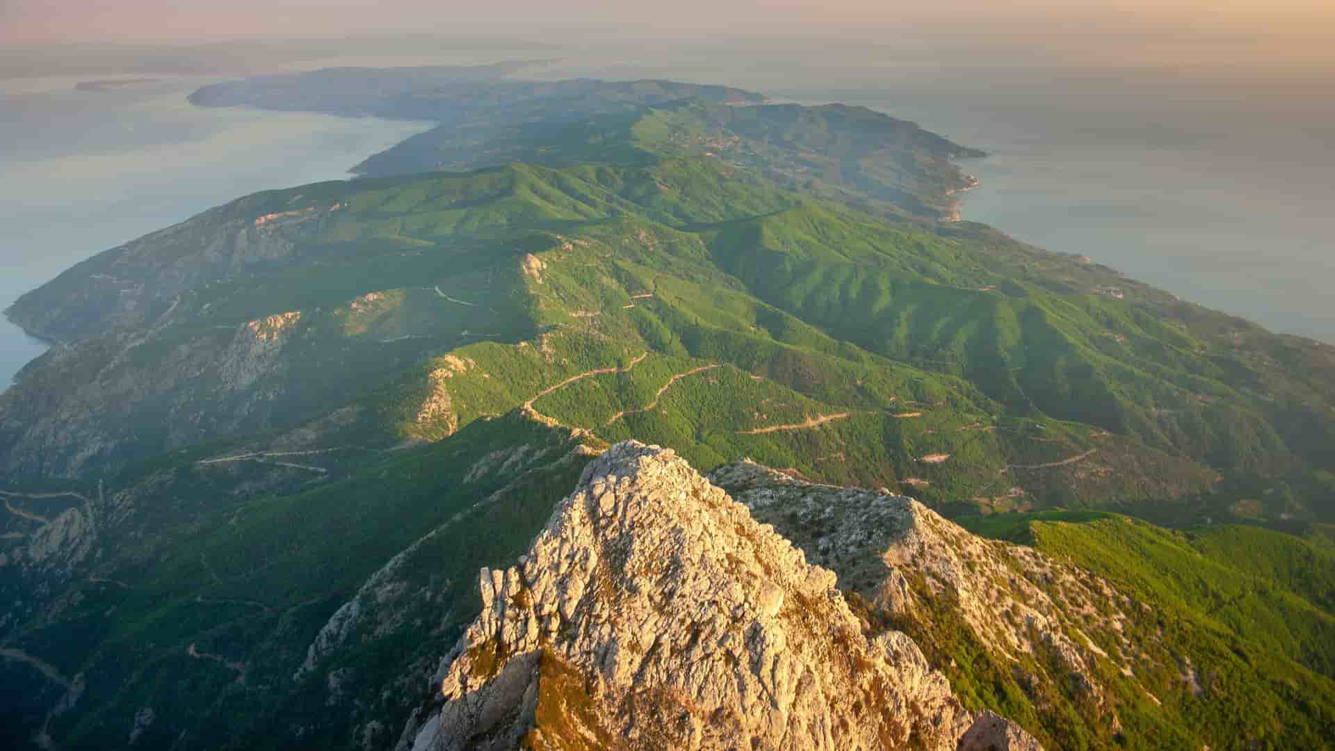 An aerial view of the rugged, green peninsula of Mount Athos in Greece, with its rocky peak rising above the sea and a winding road visible on the hillside.