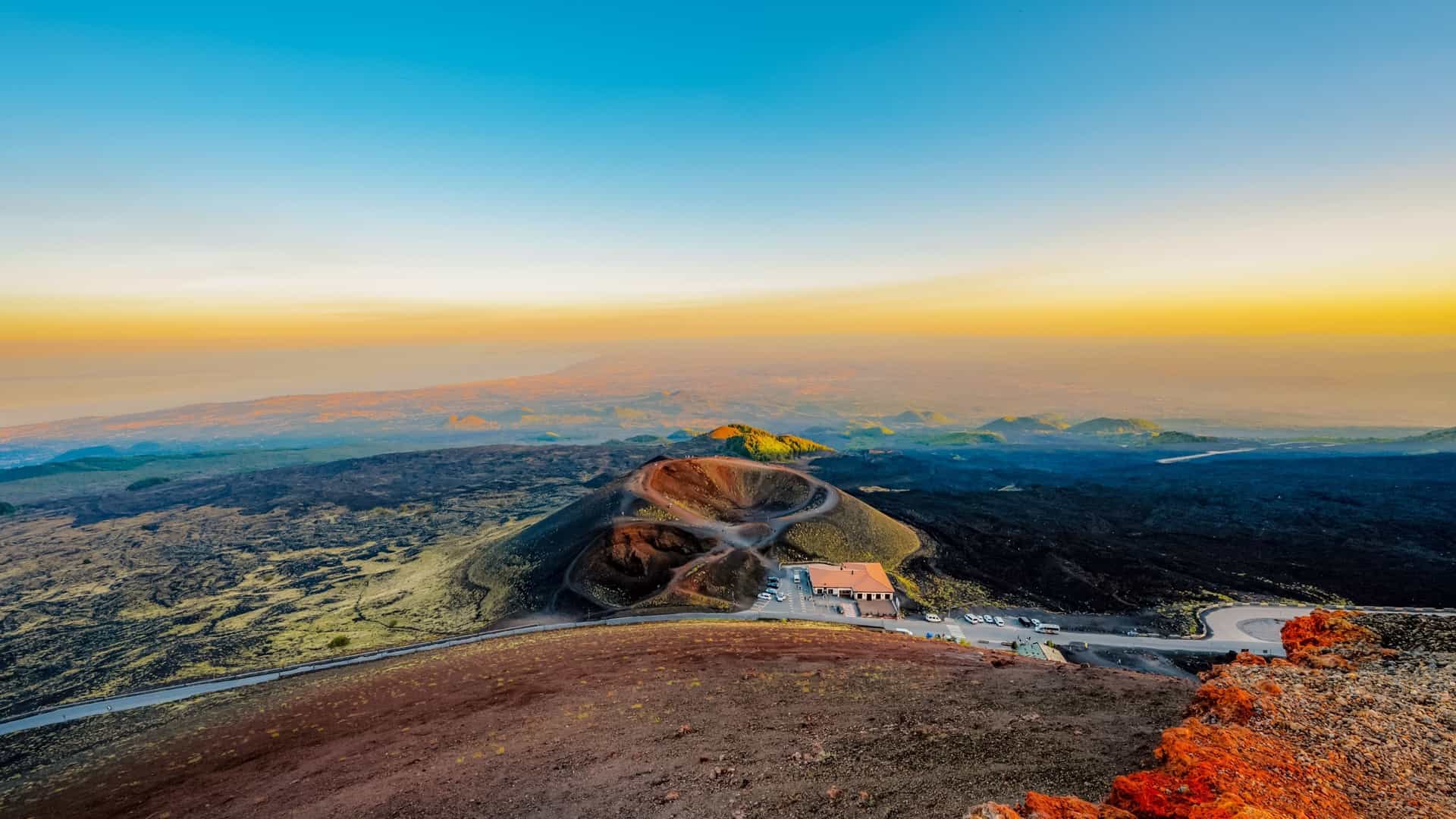 A high-angle view of Mount Etna, Italy, shows a visitor center on a road, surrounded by dark volcanic ash and rock, at sunset.