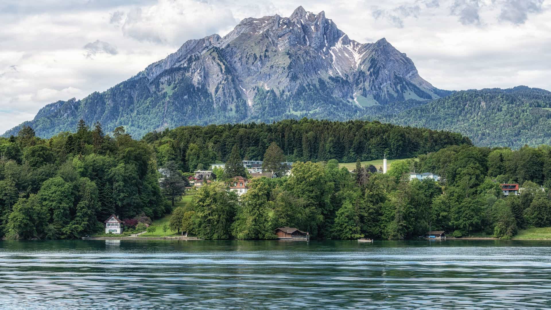 A majestic view of the rugged, snow-dusted peak of Mount Pilatus in the Swiss Alps, with a tranquil lake and a lush green forest at its base.