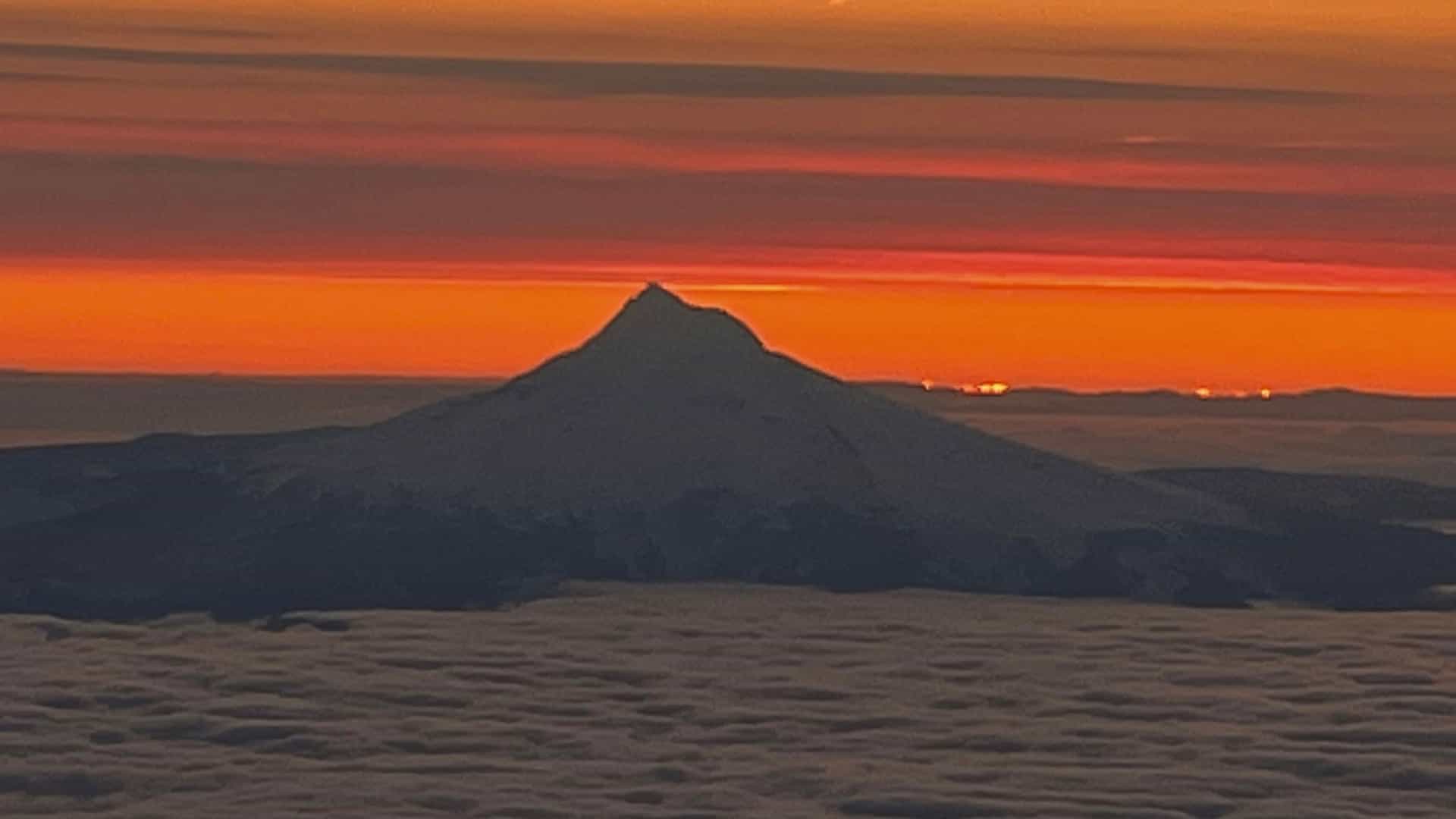 A dramatic sunset view over a cloud layer with a mountain peak silhouetted against a vibrant orange and red sky, possibly Mount Hood or Mount Rainier.