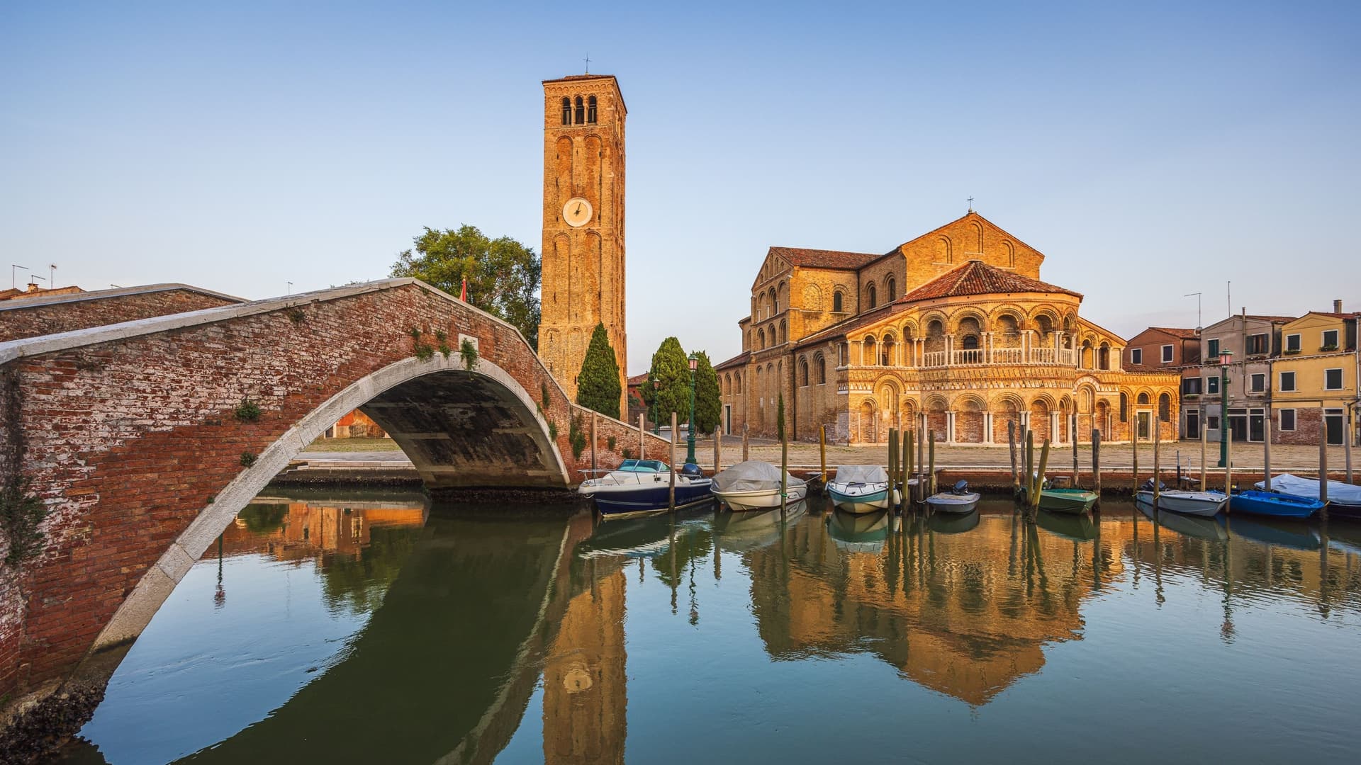 A brick bridge and a tall clock tower, reflecting in a canal, stand next to the historic Church of Santa Maria e San Donato.
