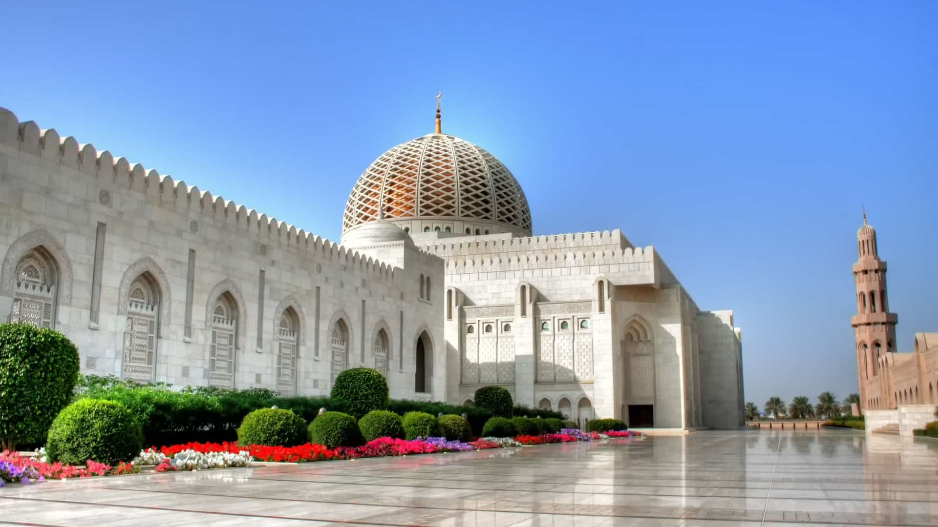 A side view of the grand Sultan Qaboos Grand Mosque in Muscat, Oman, featuring its majestic dome and minaret under a clear blue sky.