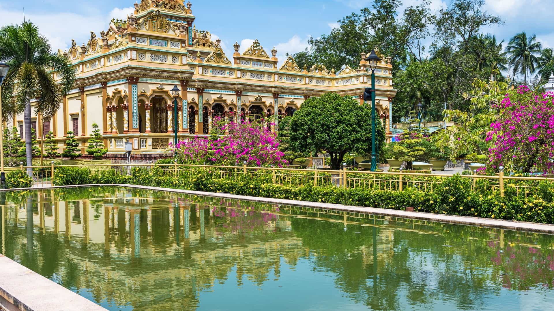 A colorful, ornate temple in My Tho, Vietnam, with intricate architectural details, surrounded by lush gardens, palm trees, and a tranquil reflecting pond.