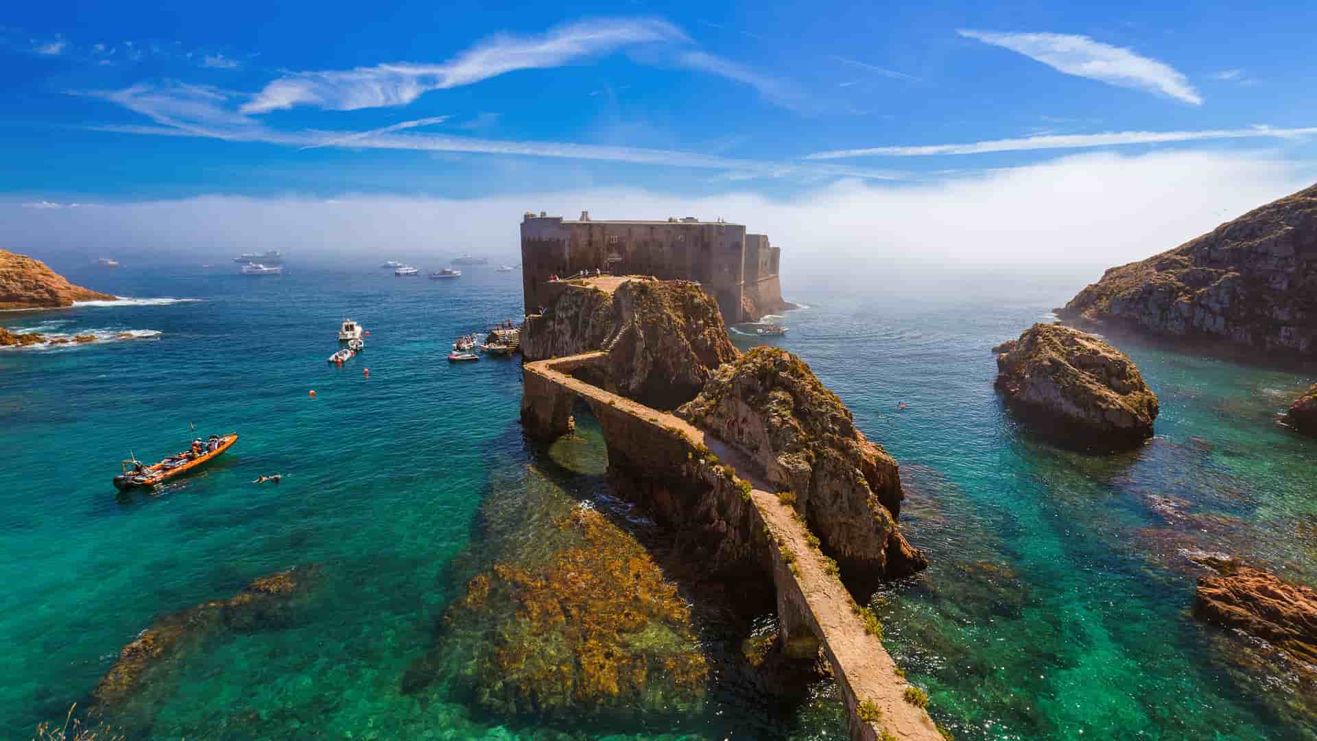 A scenic view of Forte de São João Baptista, a fortress built on a rocky islet in the Berlengas Archipelago, connected by a stone causeway over turquoise water.