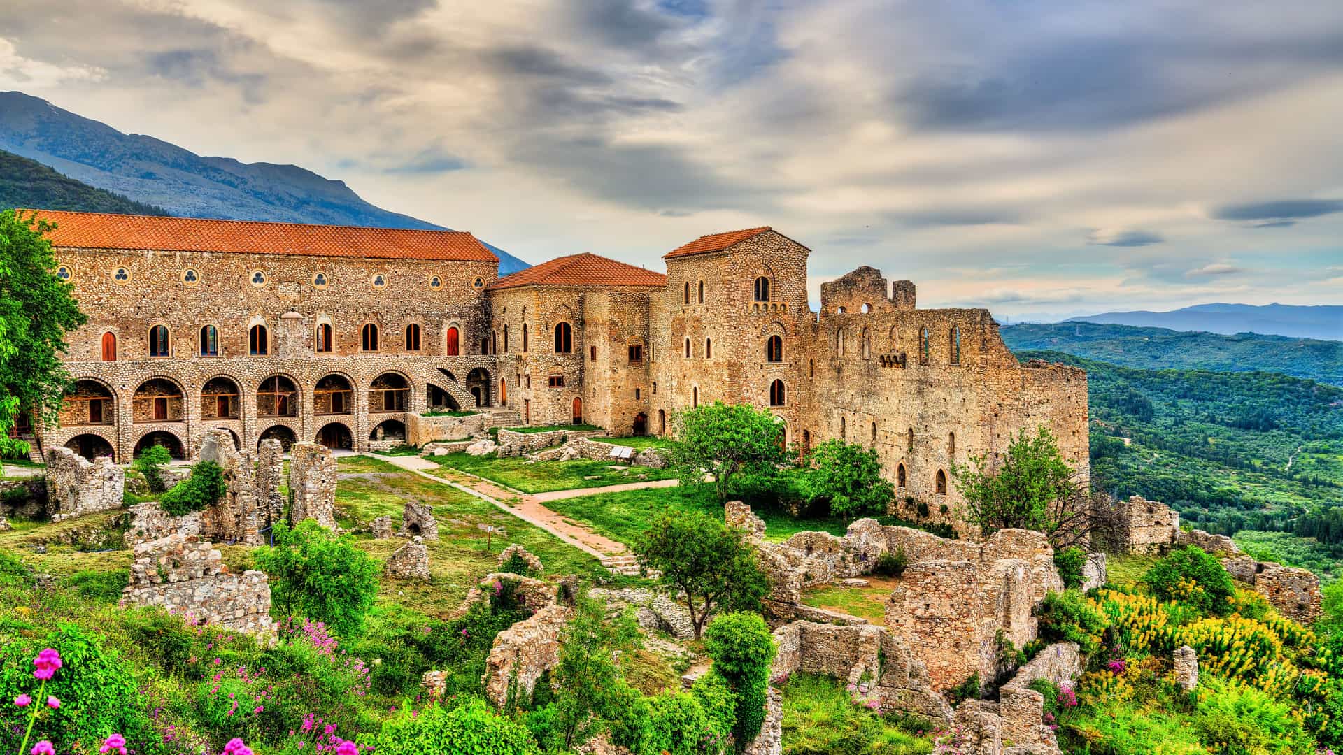"A high-angle view of the medieval city of Mystras, Greece, showcasing the remains of the Palace of the Despots and other stone buildings on a hillside with the Taygetos mountains in the background. "