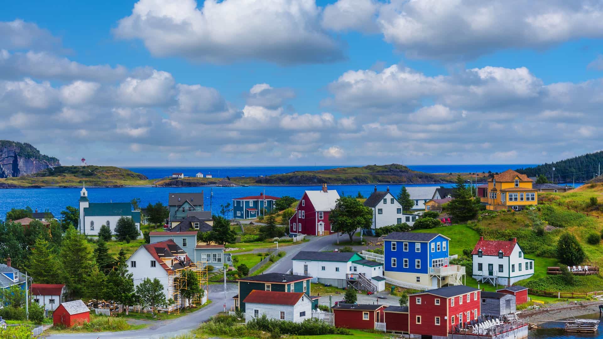 A scenic photo of a vibrant coastal town with colorful houses on a green hillside overlooking a deep blue harbor under a blue sky with white clouds.
