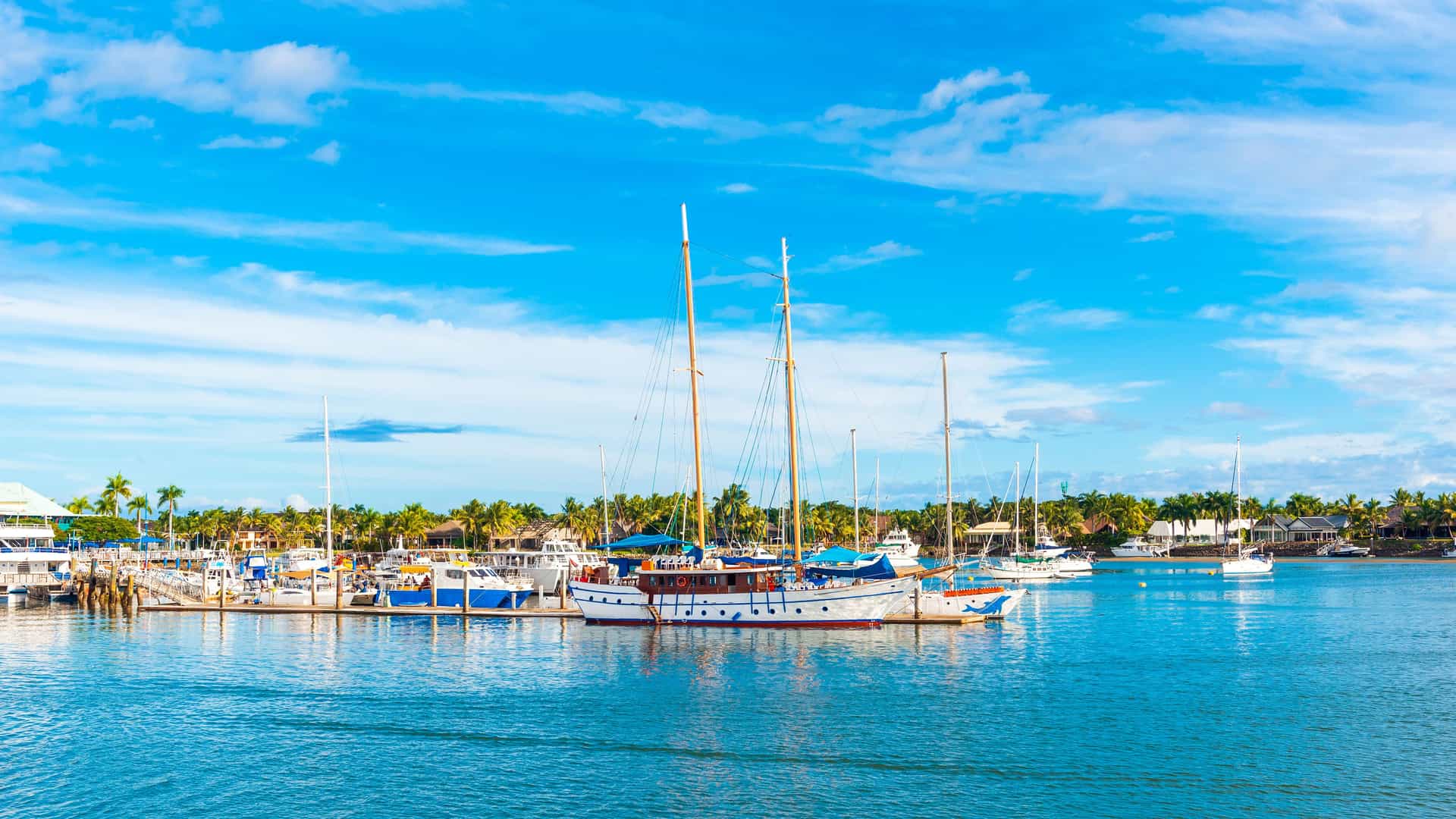 "A sunny, wide-angle shot of a marina in Nadi, Fiji, with several boats docked in the calm turquoise water under a bright blue sky with scattered clouds. "