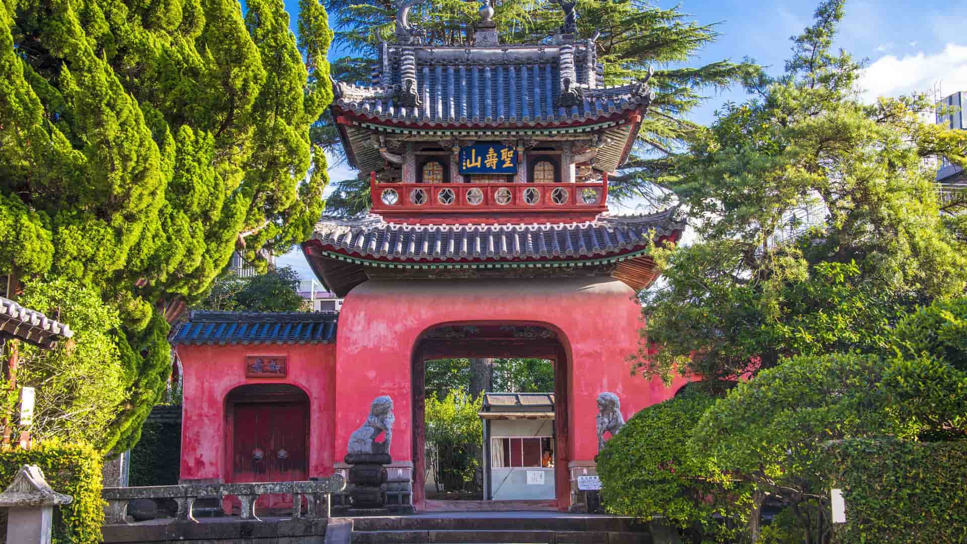 A detailed view of the vibrant, red and blue gate of Sofuku-ji Temple in Nagasaki, Japan, featuring traditional architecture and lion statues amidst lush green trees.