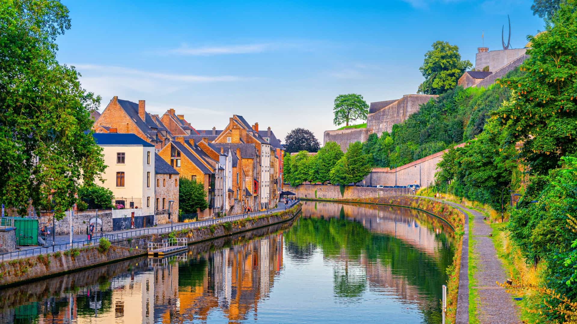 A serene canal in Namur, Belgium, with historic brick buildings reflecting in the calm water and the impressive stone walls of the citadel on a high bluff.