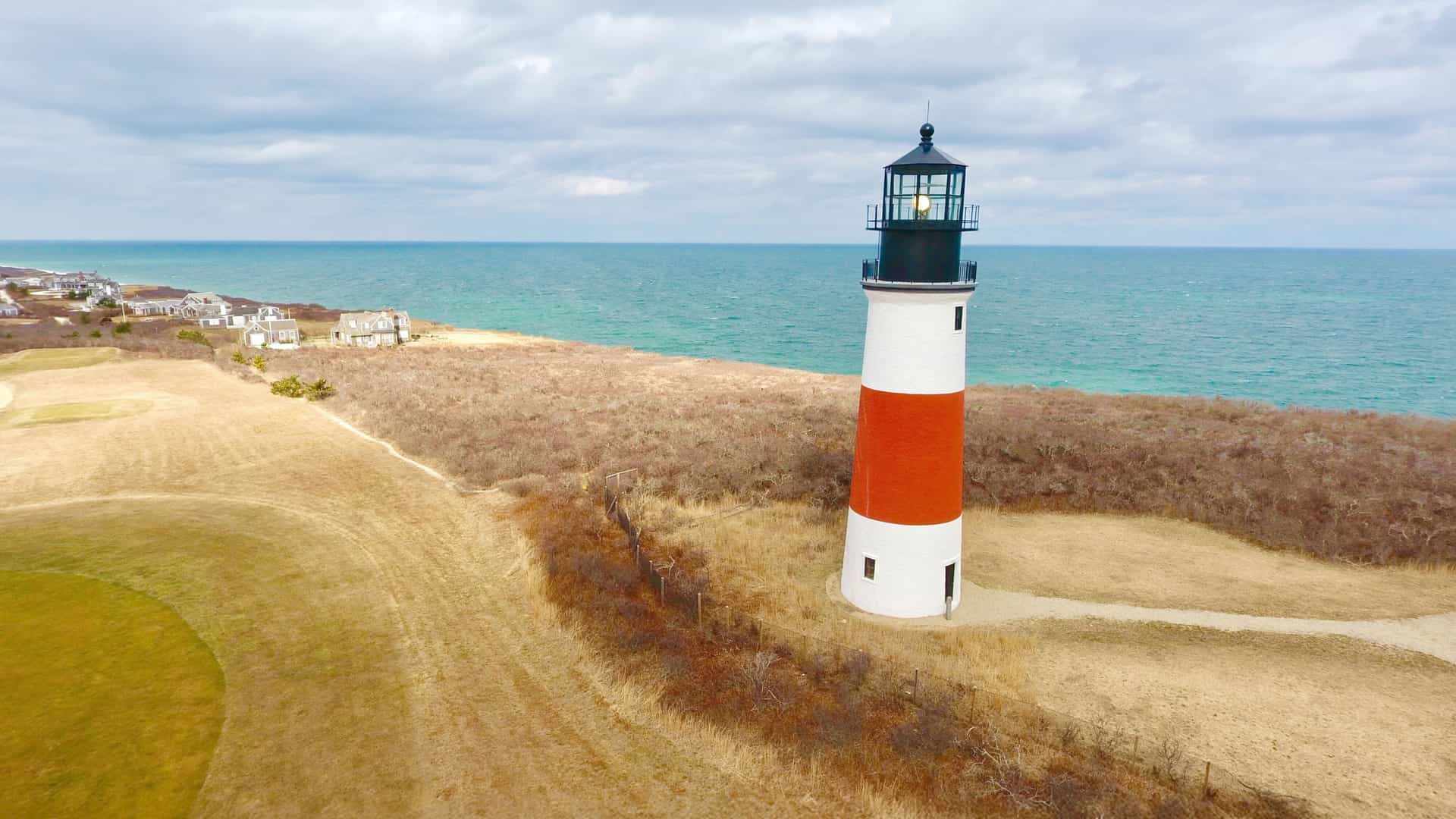 The iconic Sankaty Head Lighthouse on Nantucket, with red and white bands, stands on a grassy bluff overlooking the turquoise ocean.