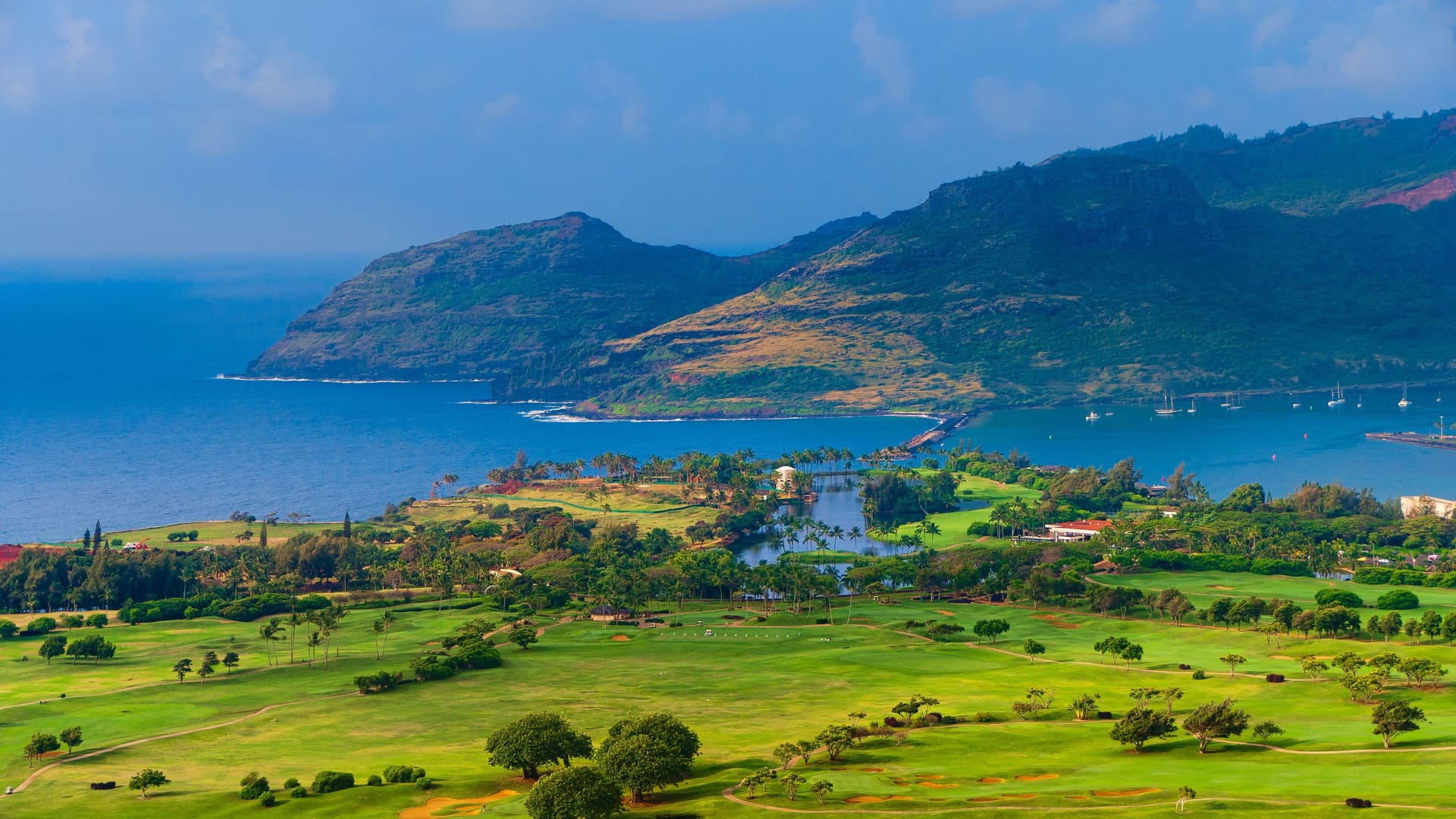 A panoramic view of a lush green golf course and resort area near Nawiliwili Harbor in Kauai, Hawaii, with the ocean and volcanic mountains in the background.