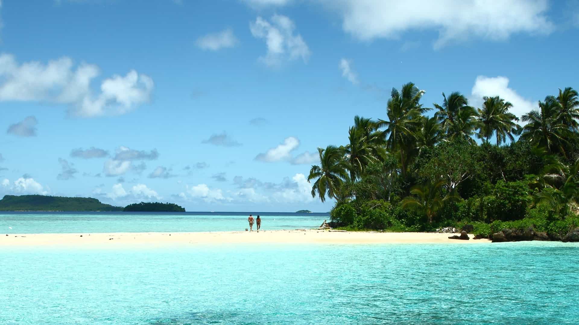 A stunning view of a pristine beach on a remote island near Neiafu, Tonga, with two people walking along the white sand, and palm trees and lush vegetation in the background.