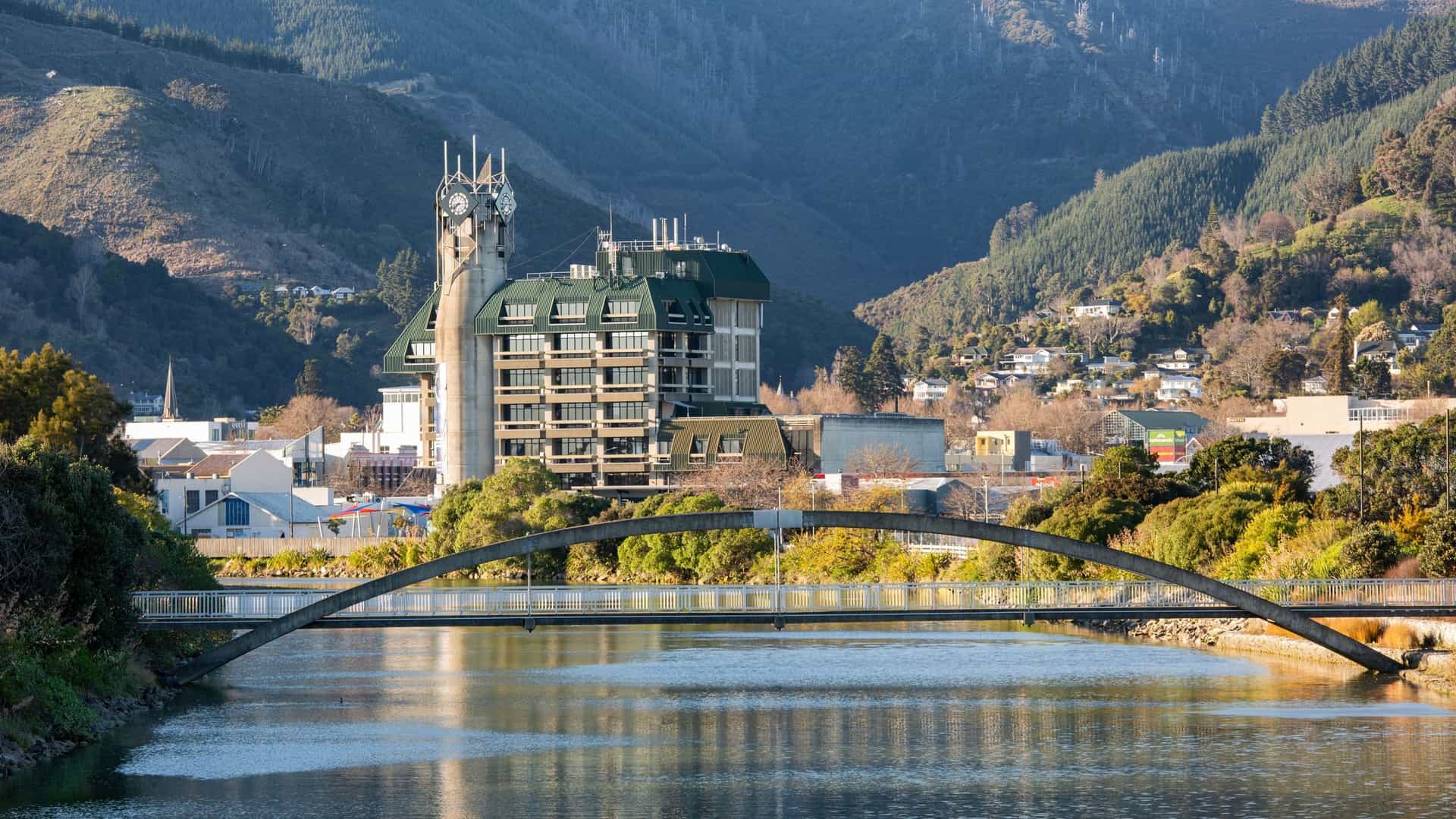 A view of Nelson, New Zealand, with a modern building and a clock tower, a pedestrian bridge over the Maitai River, and homes on the hills in the background.